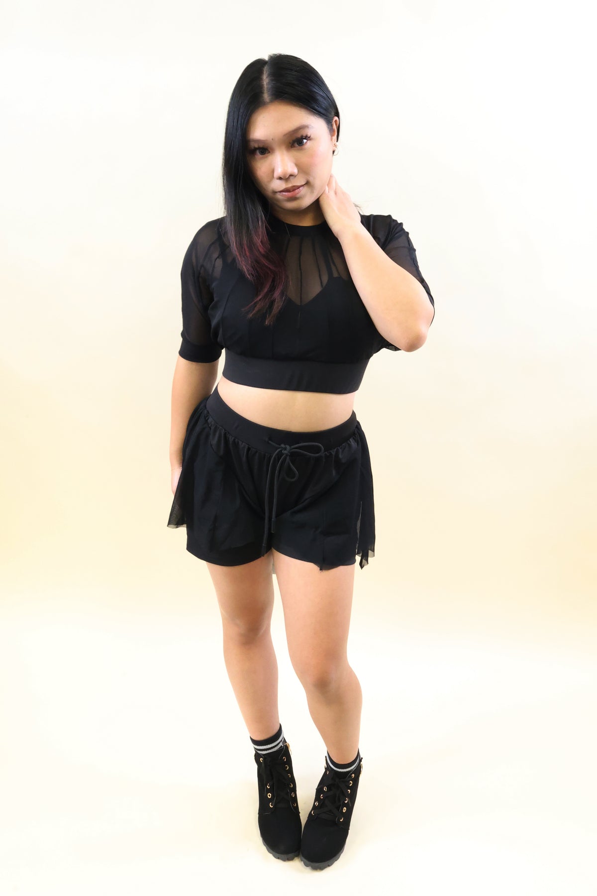 A woman modeling a black two-piece outfit consisting of a sheer short-sleeve crop top and matching black shorts with a drawstring, paired with black lace-up ankle boots, against a plain cream background.