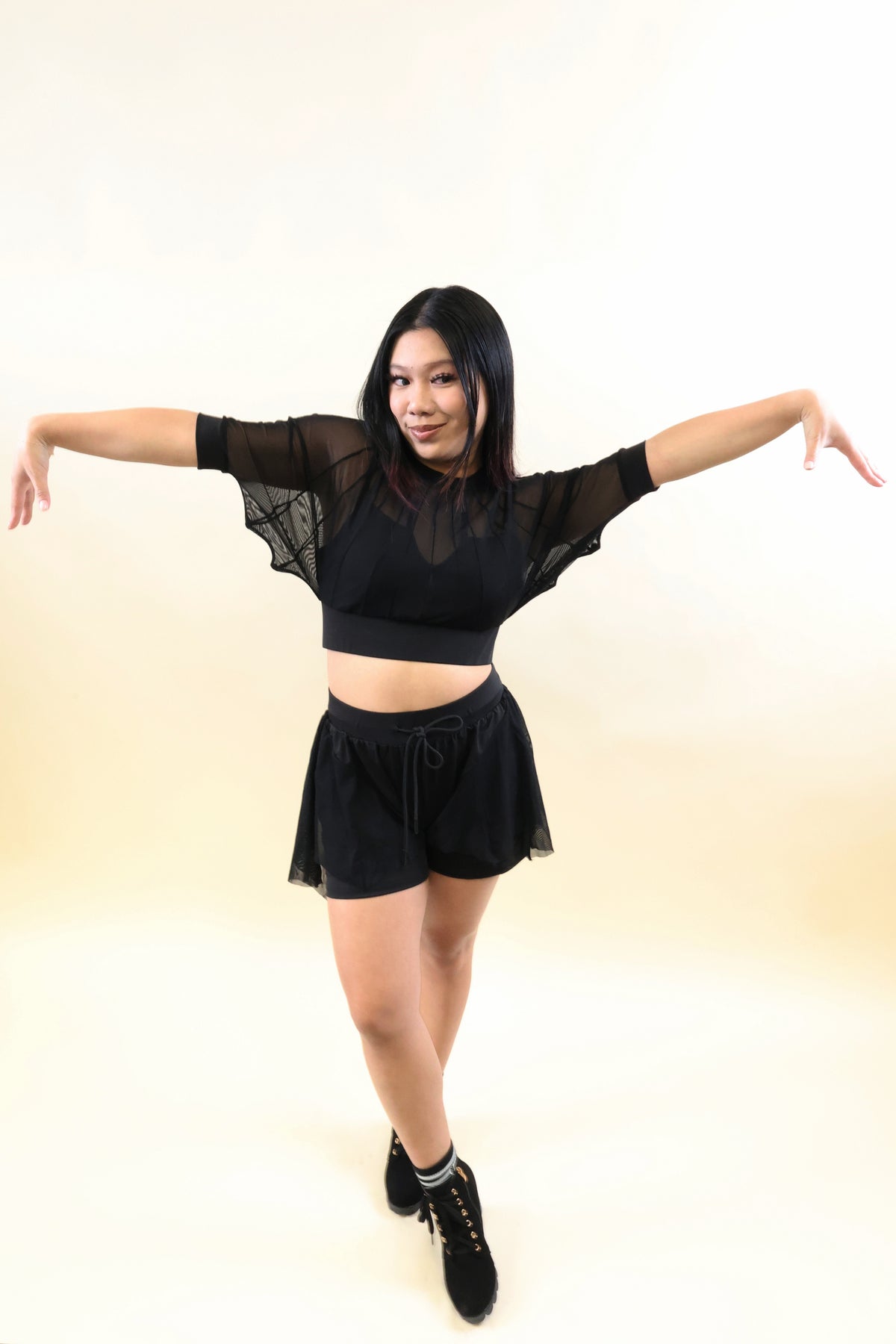 A woman modeling a black two-piece outfit consisting of a sheer long-sleeve crop top with mesh flutter sleeves and matching black shorts, posed against a plain cream-colored studio background.