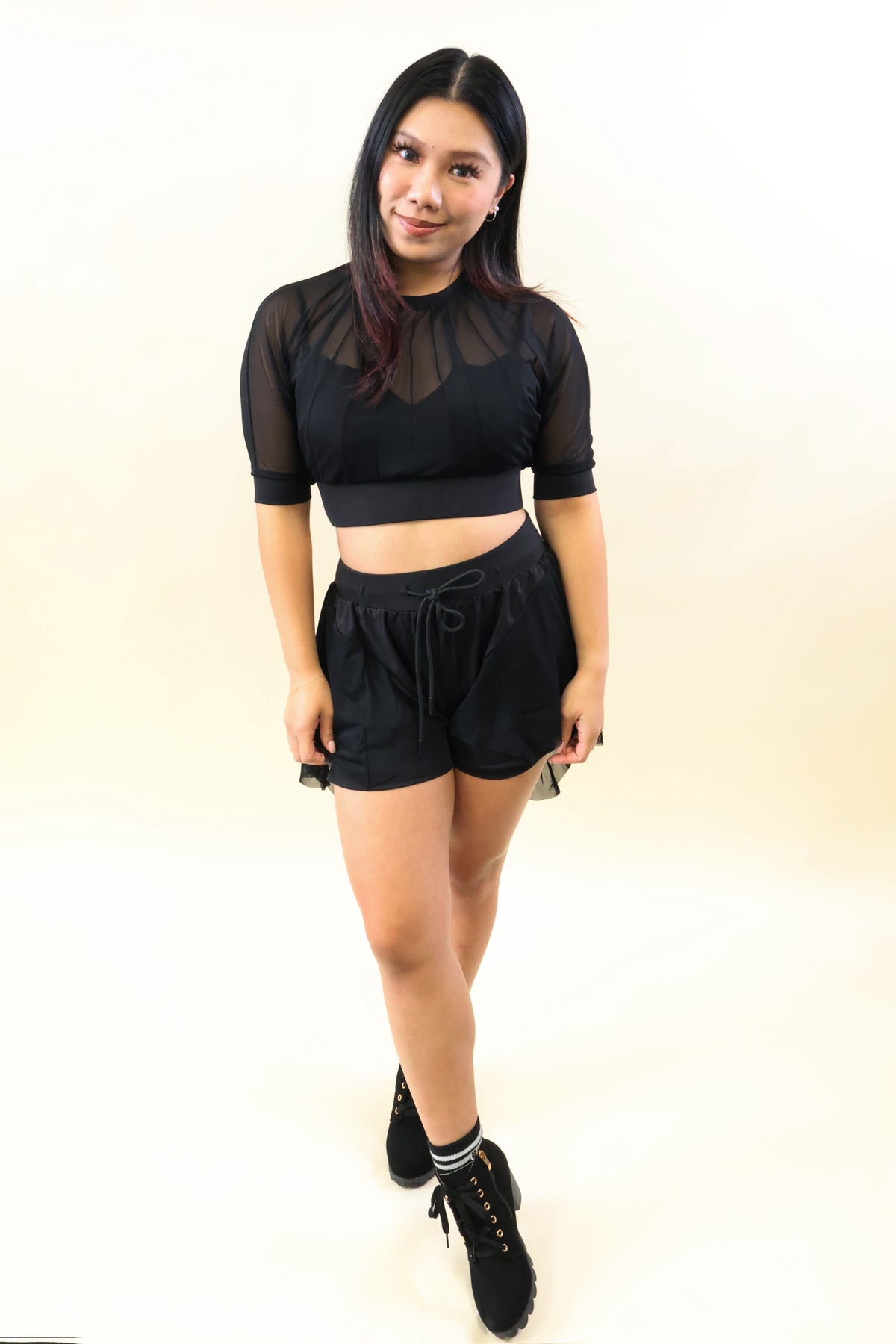 A woman modeling a black two-piece outfit consisting of a sheer short-sleeve crop top with a mesh yoke and black drawstring shorts, paired with black lace-up boots, against a plain off-white studio background.