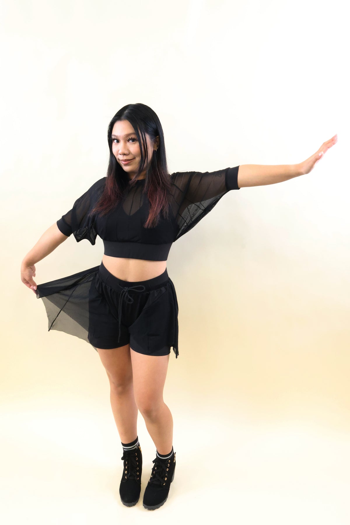 A woman modeling a black two-piece outfit consisting of a sheer short-sleeve crop top and matching black shorts, paired with black lace-up boots, against a plain cream-colored studio background.