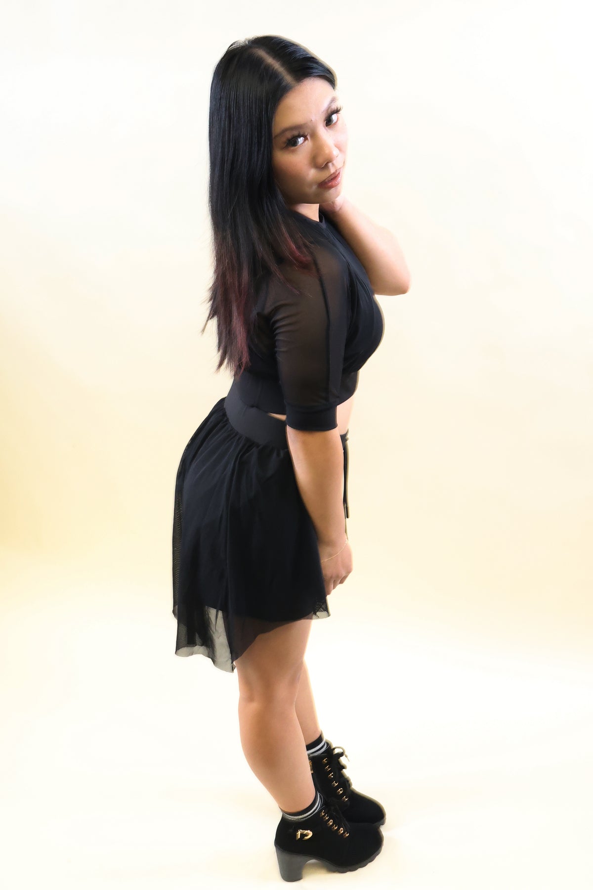 A woman posing in a black sheer long-sleeve top and matching black mini skirt with a waist tie, paired with black platform boots, against a plain cream studio background.