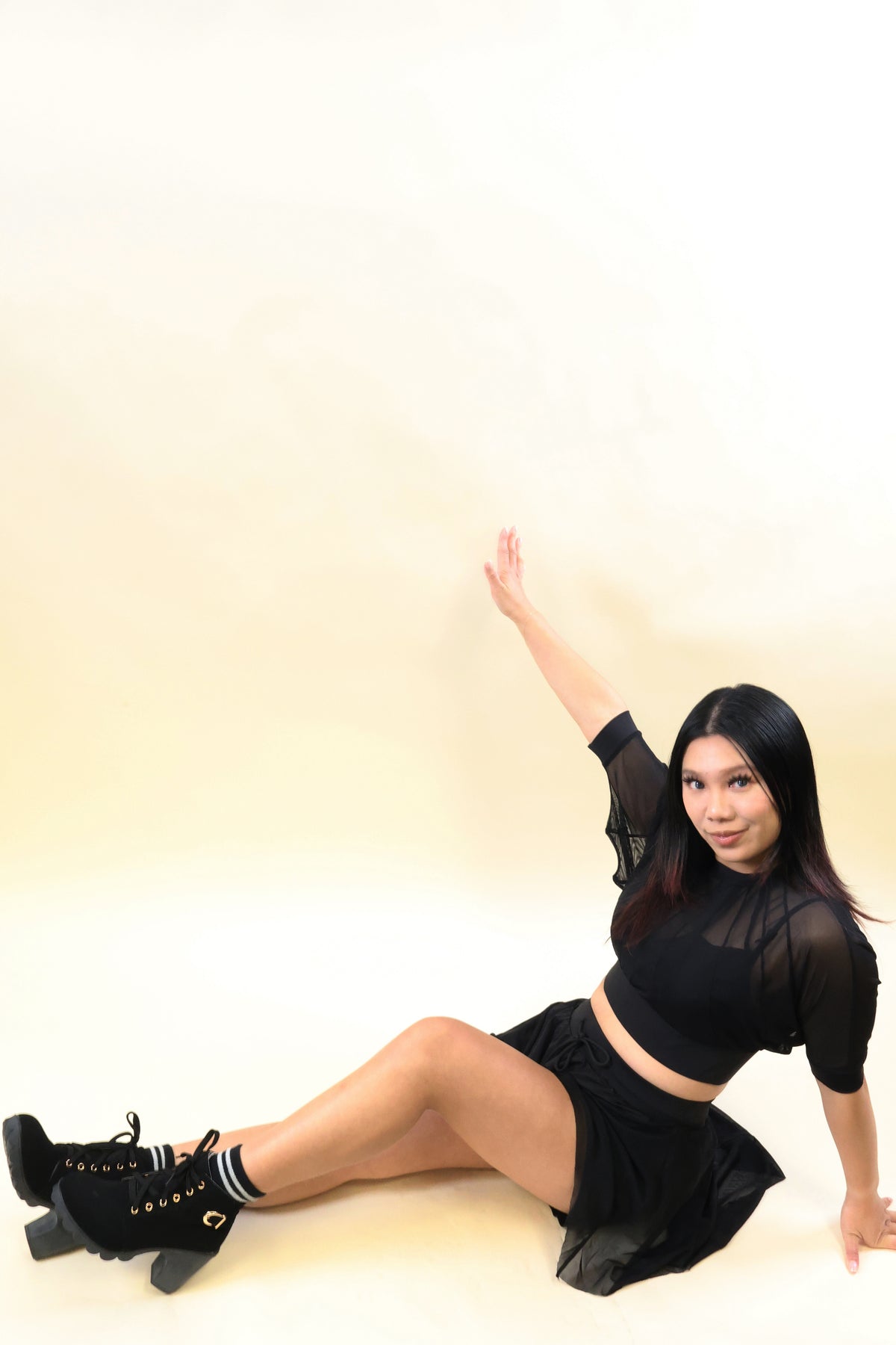 A woman wearing a black sheer long-sleeve crop top, black mini skirt, and black lace-up ankle boots poses on a light beige studio floor against a matching beige background.