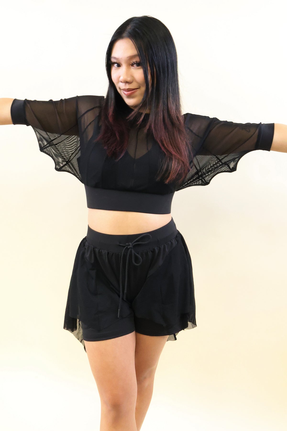 A woman modeling a black two-piece activewear set consisting of a sheer long-sleeve crop top with wide mesh sleeves and matching black drawstring shorts, posed against a plain cream background.