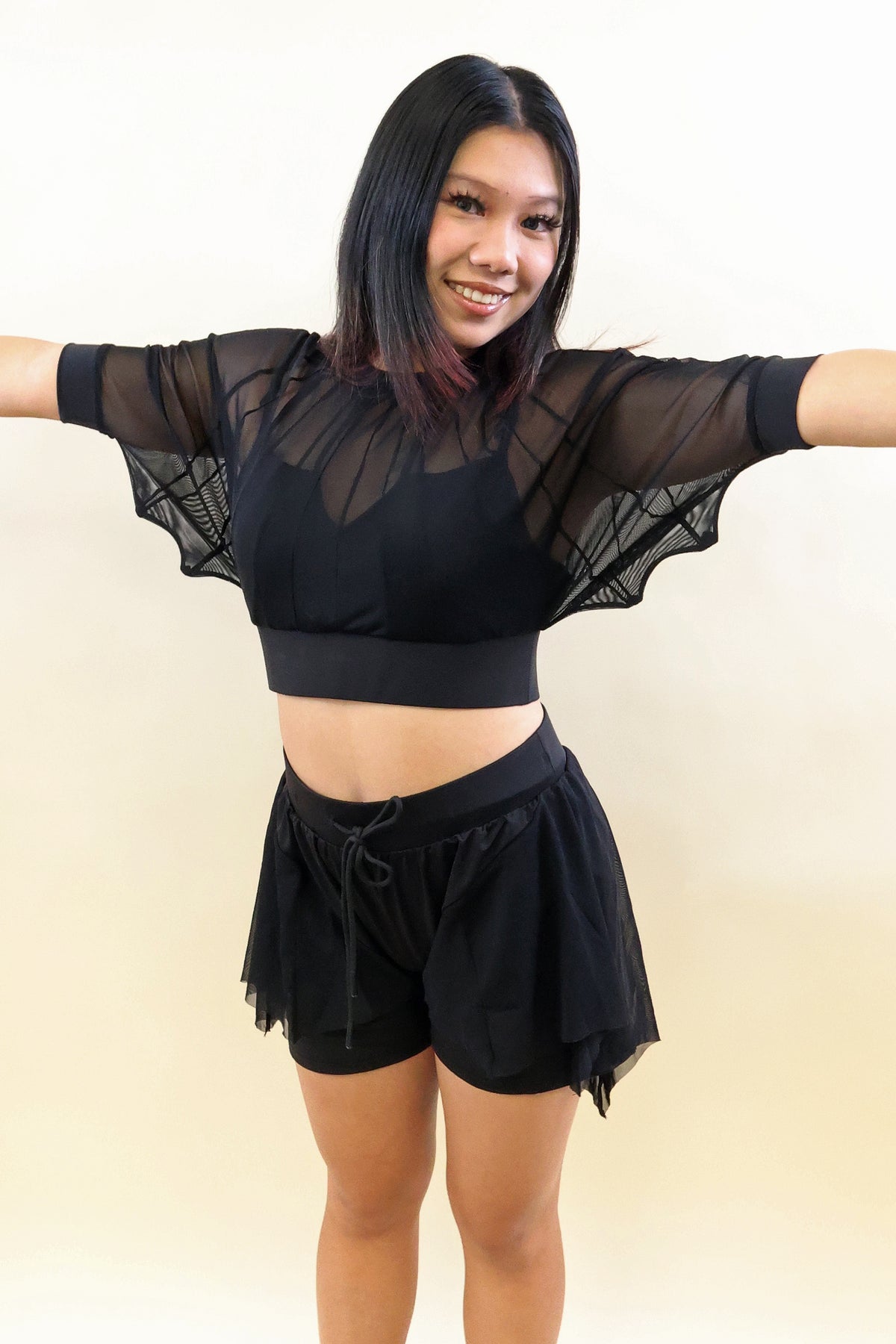 A woman modeling a black two-piece activewear set consisting of a sheer mesh short-sleeve crop top with wide flared sleeves and matching black shorts, posed against a plain light beige background.