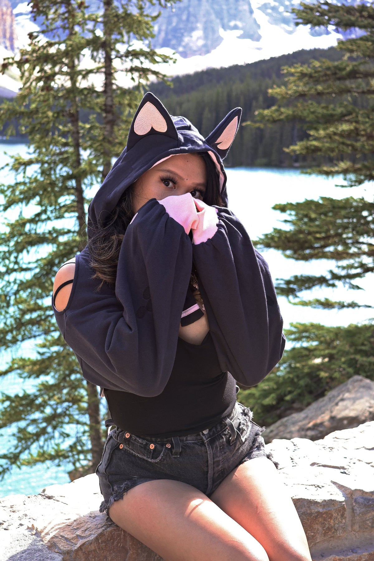 Woman modeling the Nyahallo Cat Shrug featuring cat ears, oversized sleeves, and embroidered paw print cuffs while posing beside a scenic mountain lake.