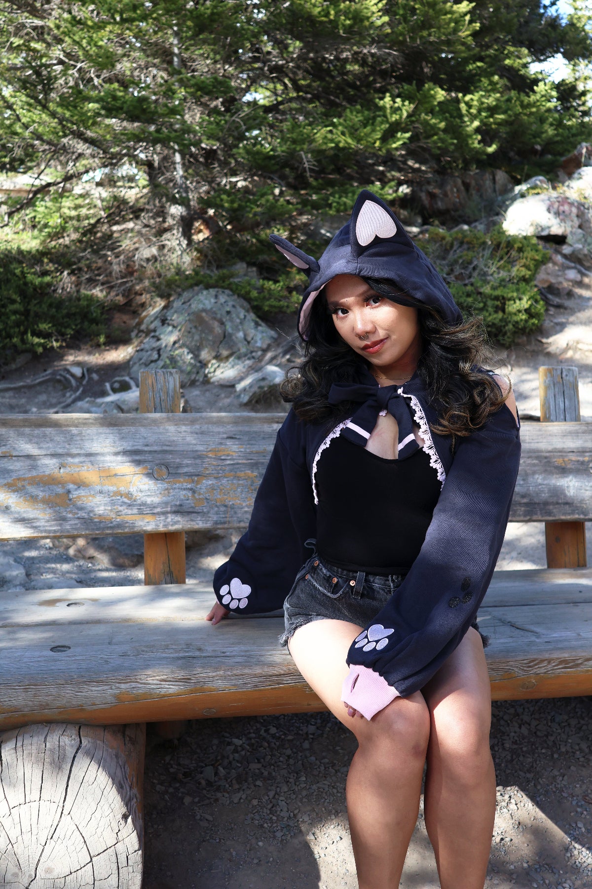 Model wearing the Nyahallo Cat Shrug with cat ears, embroidered paw print sleeves, and lace-up front while sitting on a rustic wooden bench outdoors.