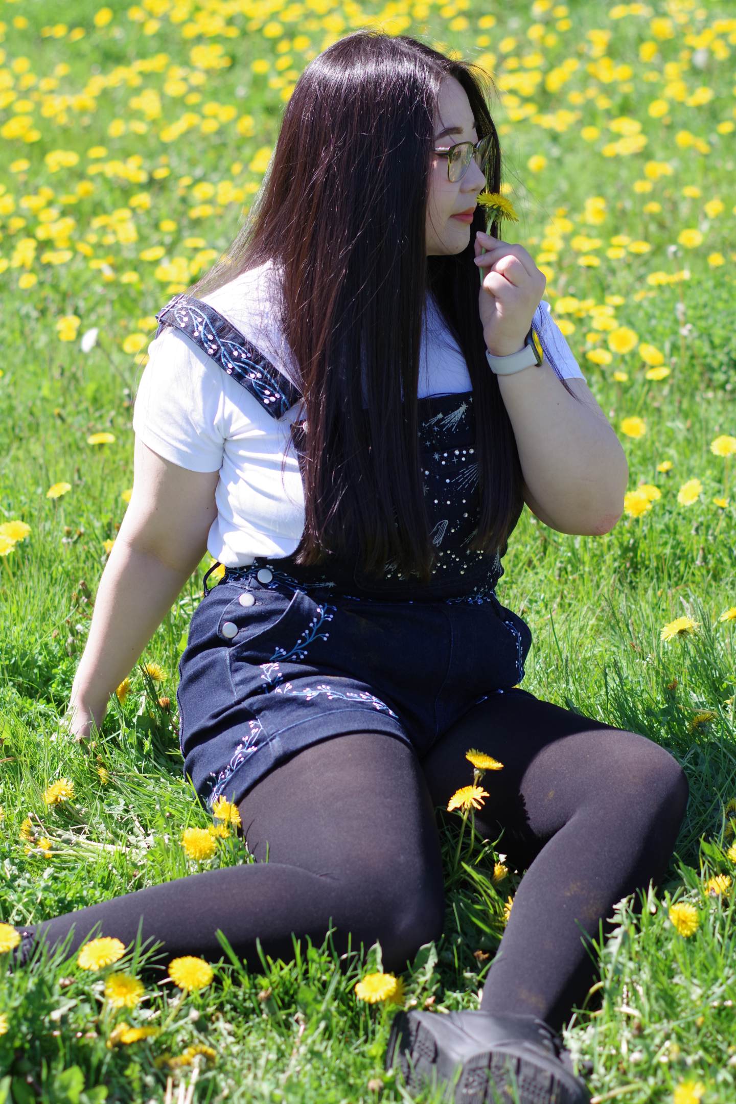 Model sitting in a field of yellow flowers wearing navy celestial embroidered overalls with constellation details layered over a white t-shirt.