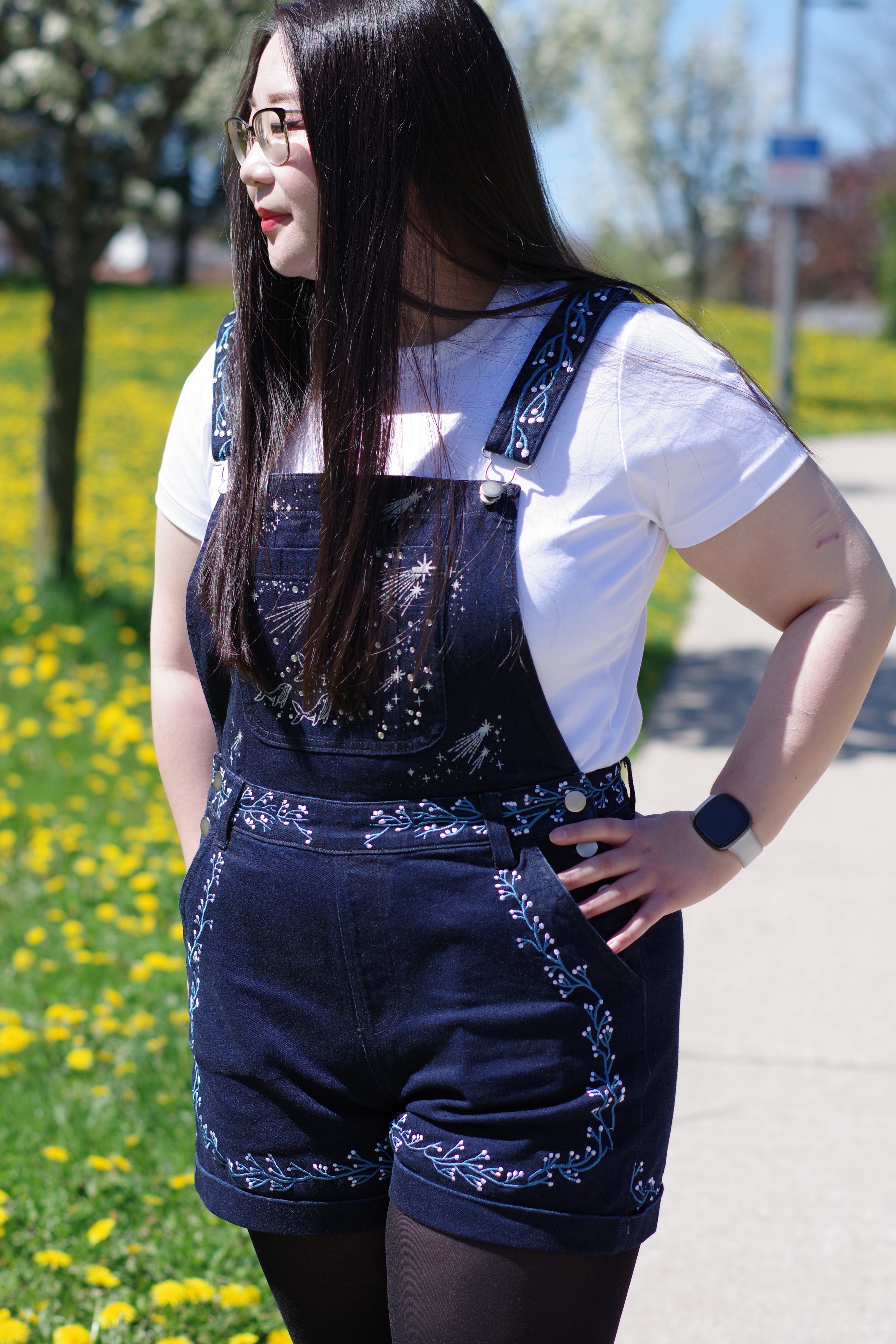Model wearing navy celestial embroidered overalls with constellation and shooting star details over a white t-shirt beside a field of yellow flowers.