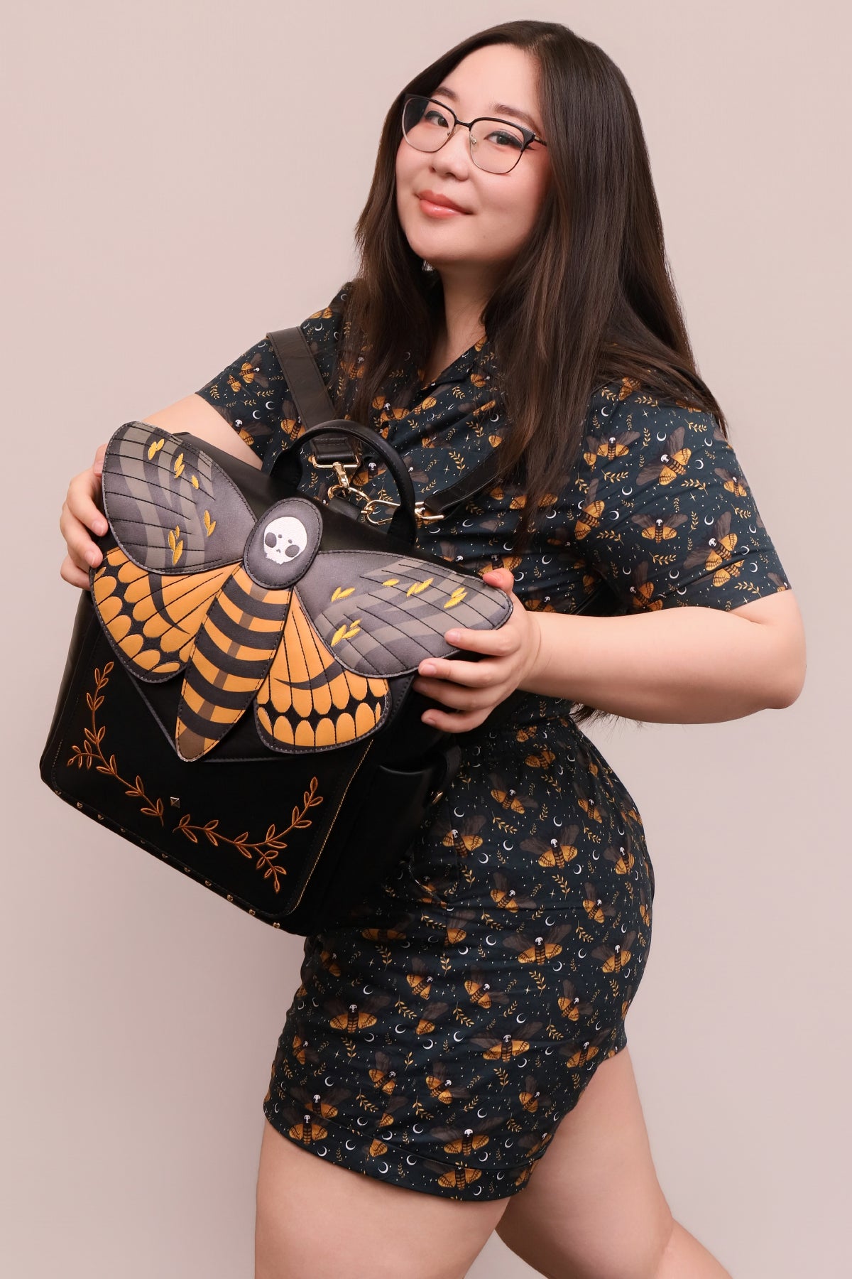 Model holding the Death's-Head Hawkmoth Backpack featuring dimensional moth wings, skull detailing, embroidered botanical accents, and gothic nature-inspired artwork.