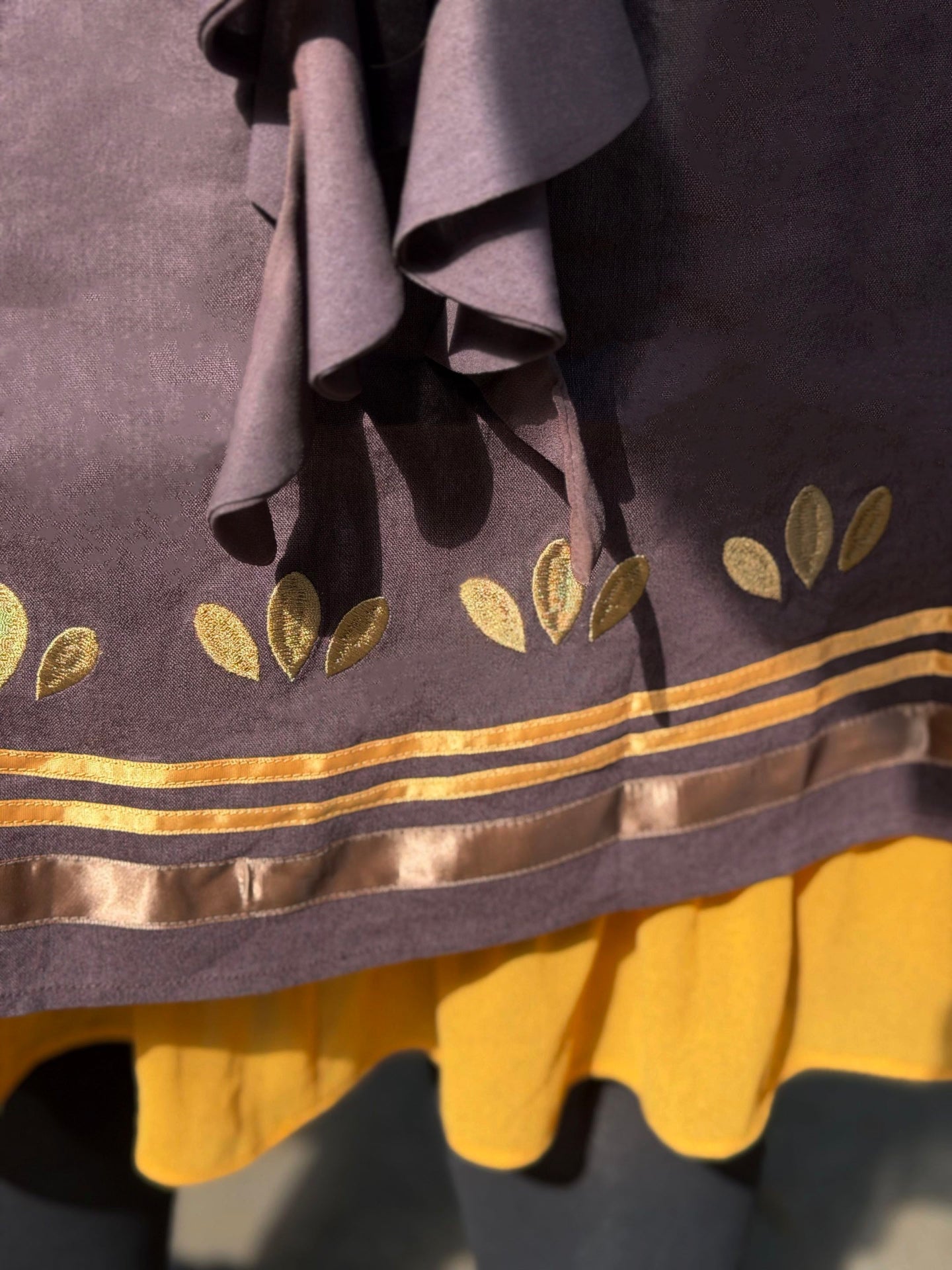Close-up detail of the Death’s-Head Hawkmoth Dress hem featuring embroidered golden leaf motifs, satin ribbon trim, and layered golden ruffles in warm sunlight.