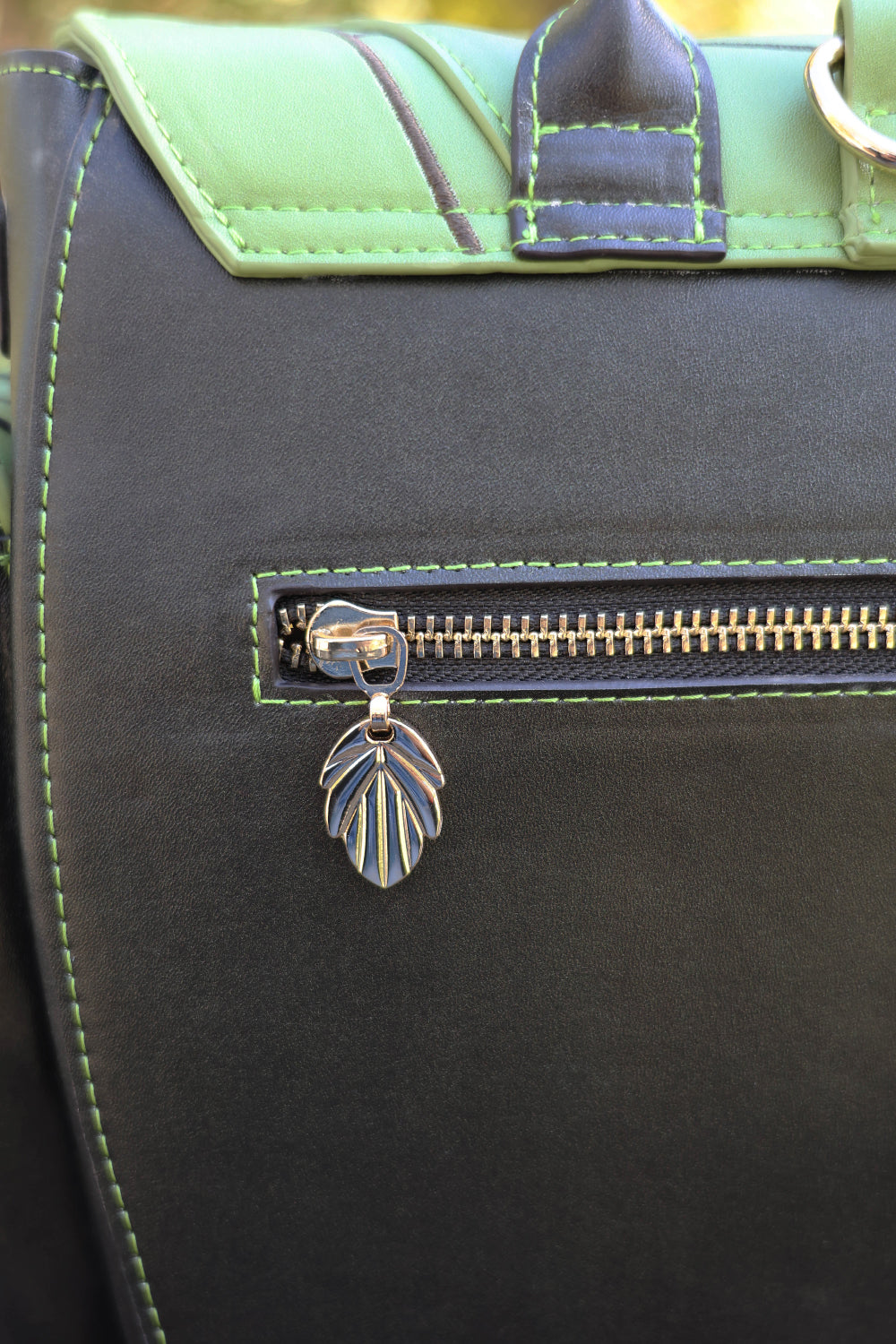 Close-up of a dark brown bag with green accents and a leaf-shaped charm on a blurred natural background