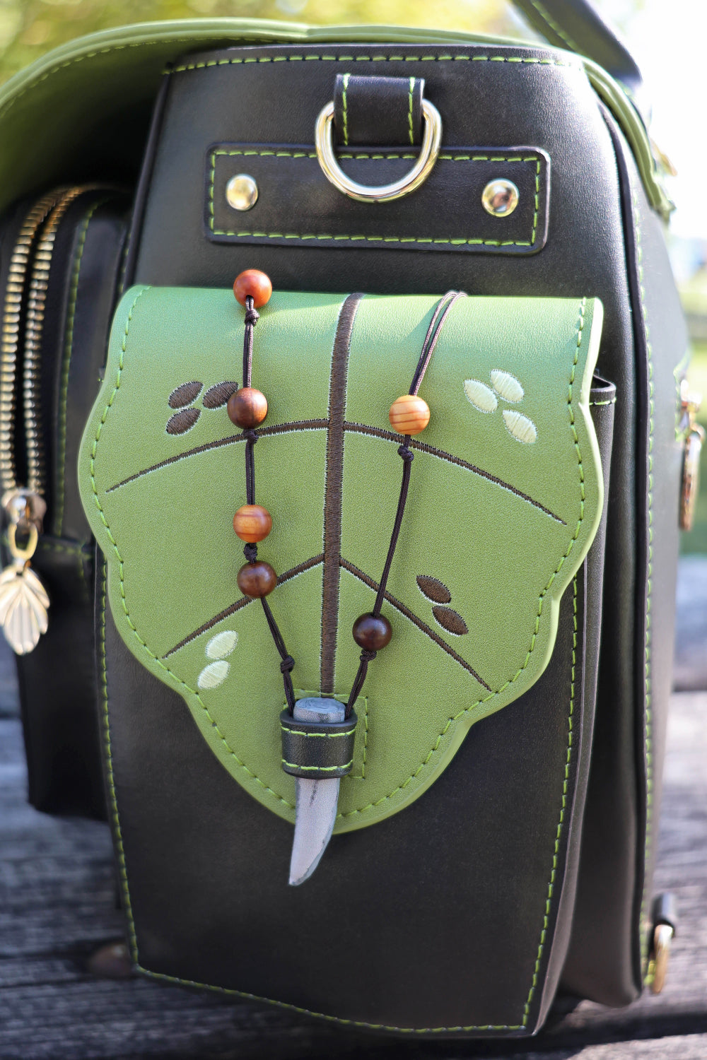 Side view of a green and dark brown pouch with decorative details, set against a blurred natural background