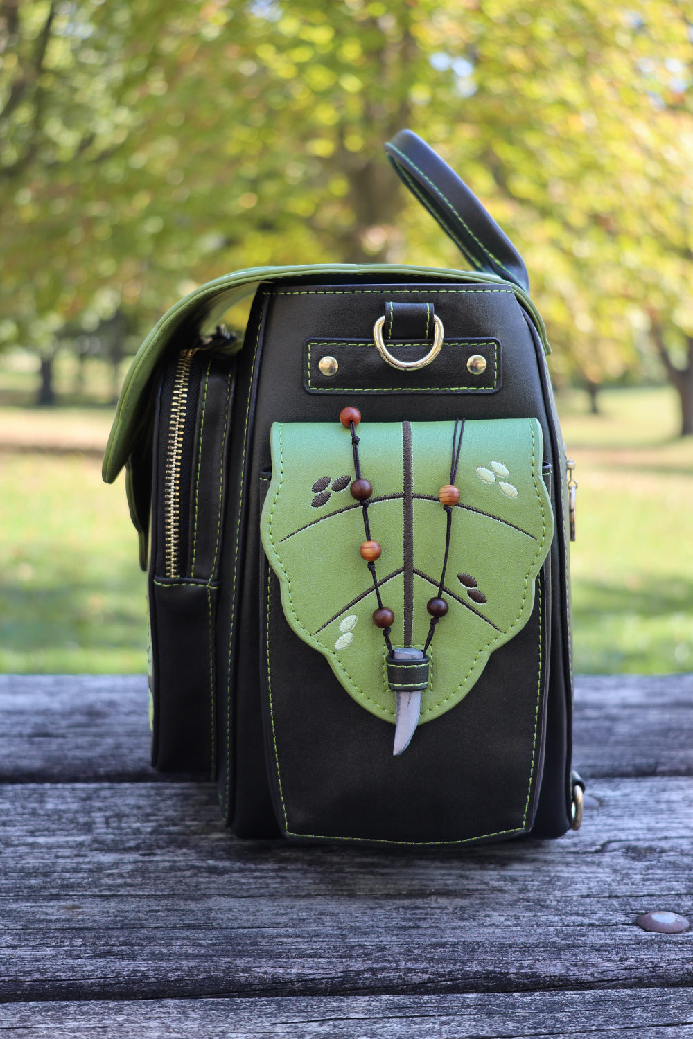 Dark brown and green handbag with green leaf design on a wooden surface with blurred greenery in the background