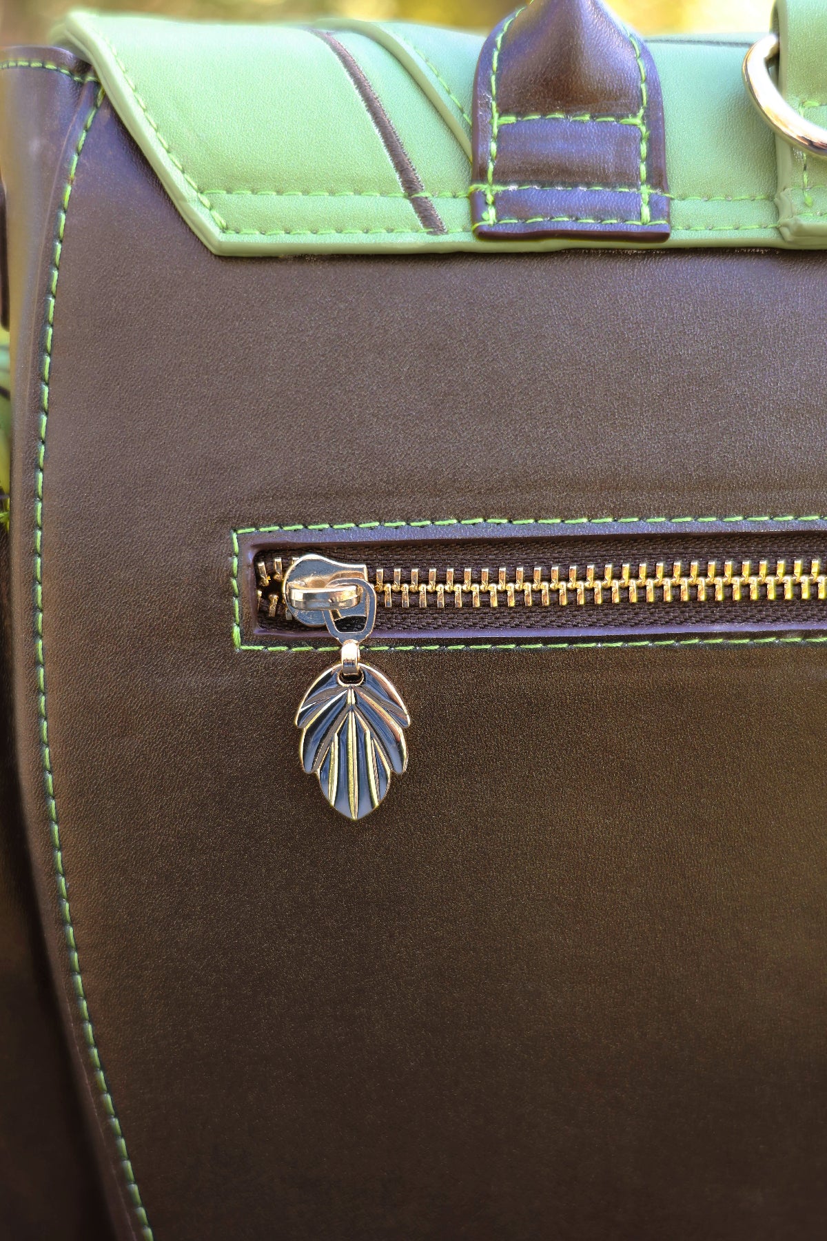 Close-up of the Druid backpack back pocket featuring brown faux leather, green contrast stitching, gold zipper hardware, and a leaf-shaped zipper pull.