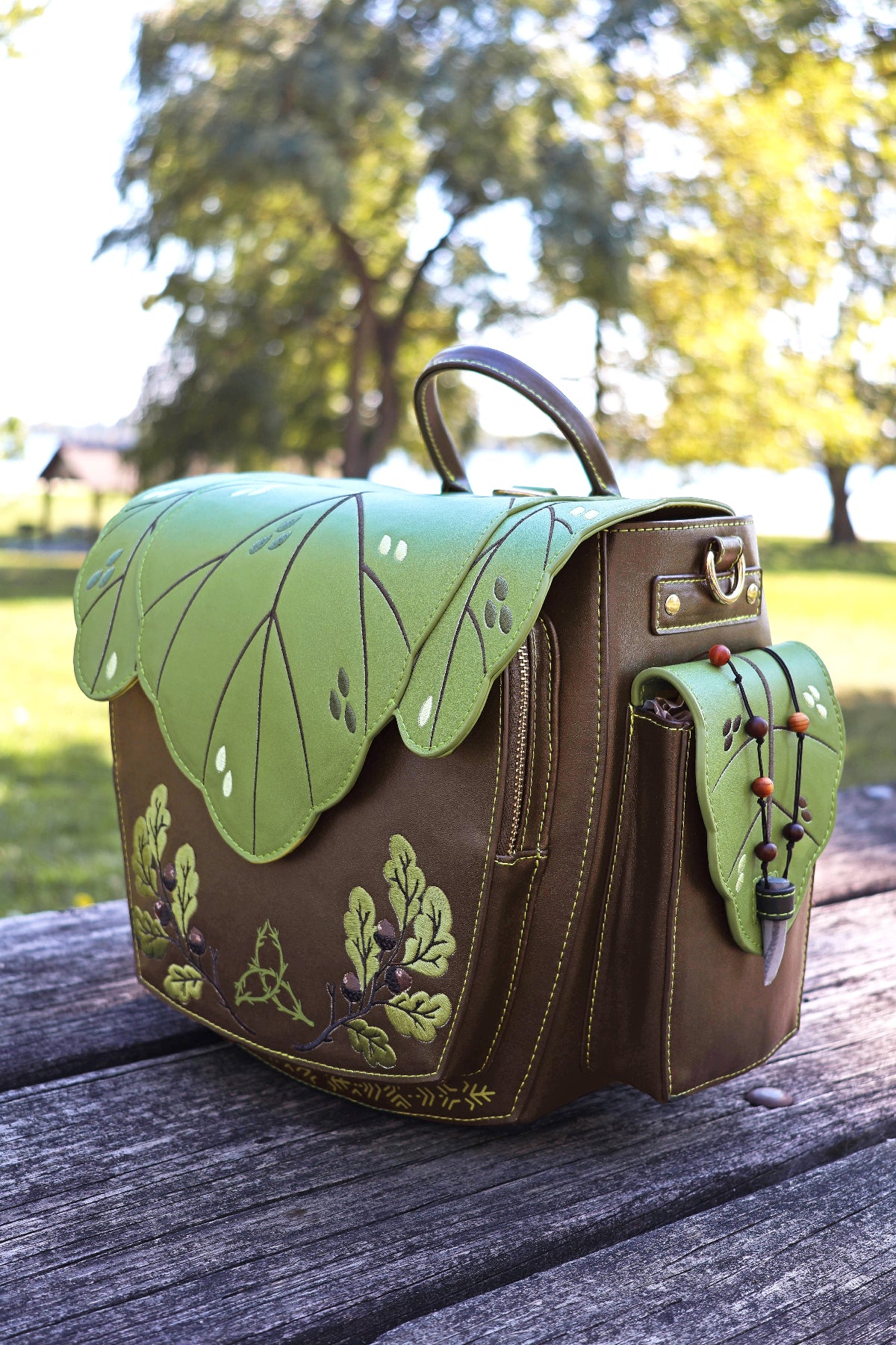 Druid leaf backpack in brown and green with an embroidered botanical design, layered leaf flap, decorative side pocket, bead details, and gold hardware.