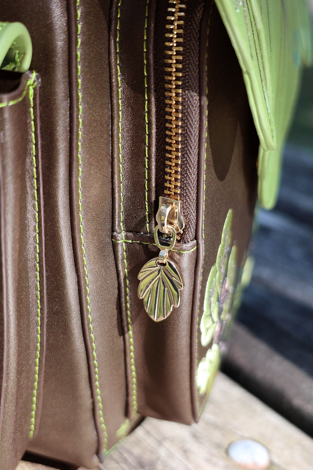 Close-up of the Druid backpack showing brown faux leather, green contrast stitching, gold zipper hardware, and a decorative leaf-shaped zipper pull.