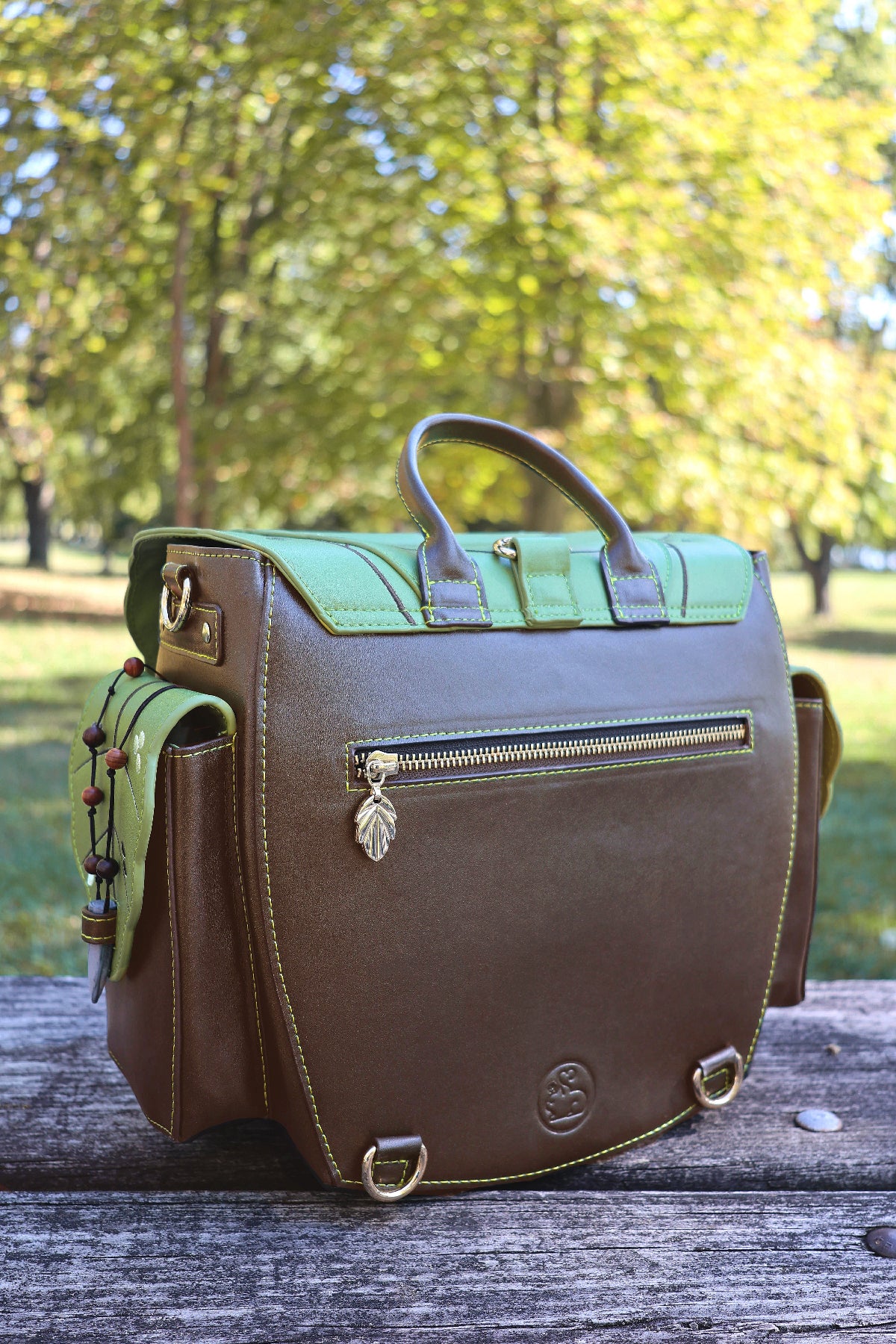Back view of the green and brown Druid backpack showing the top handle, zippered back pocket, side pockets, and gold hardware.