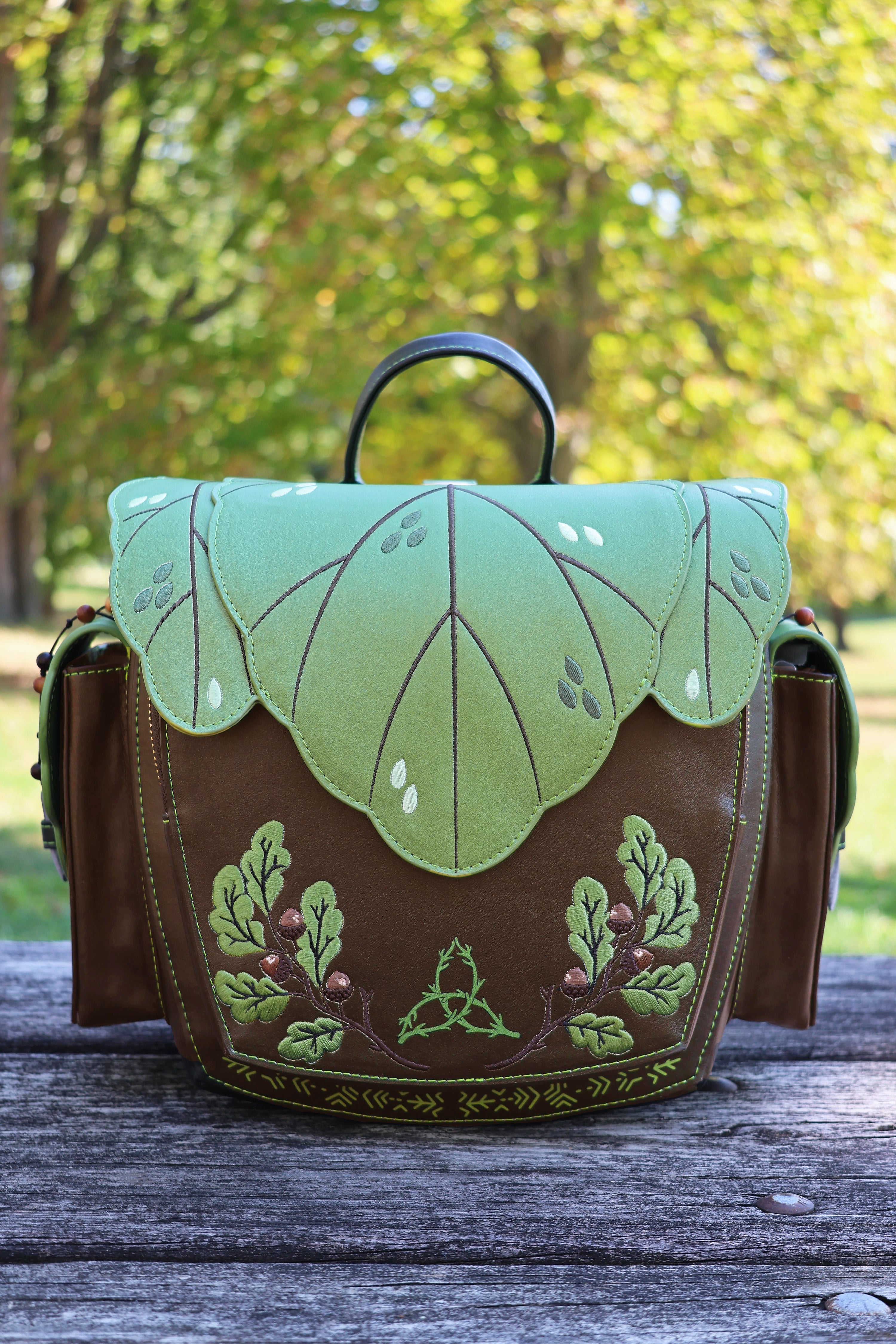 Front view of the green and brown Druid backpack featuring a layered leaf-shaped flap, woodland embroidery, and botanical details in a forest setting.
