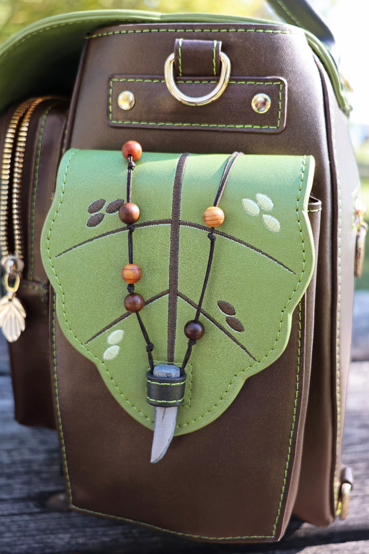 Close-up of the Druid backpack side pocket featuring a green leaf-shaped flap, embroidered details, wooden bead accents, green contrast stitching, and gold hardware.