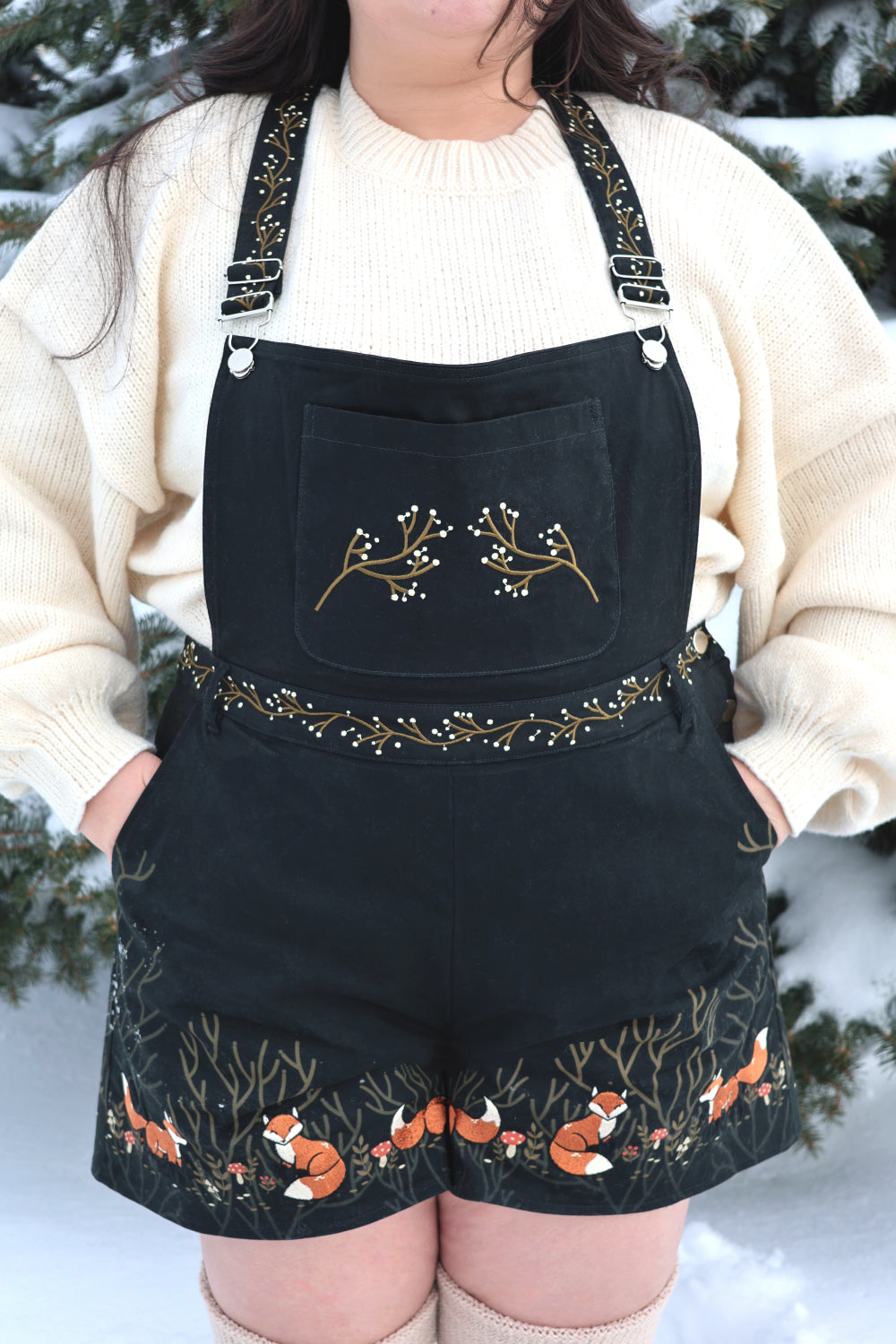 Forestcore inspired black overalls with fox embroidery, woodland vine details, and cozy cream knit styling in a snowy outdoor setting.