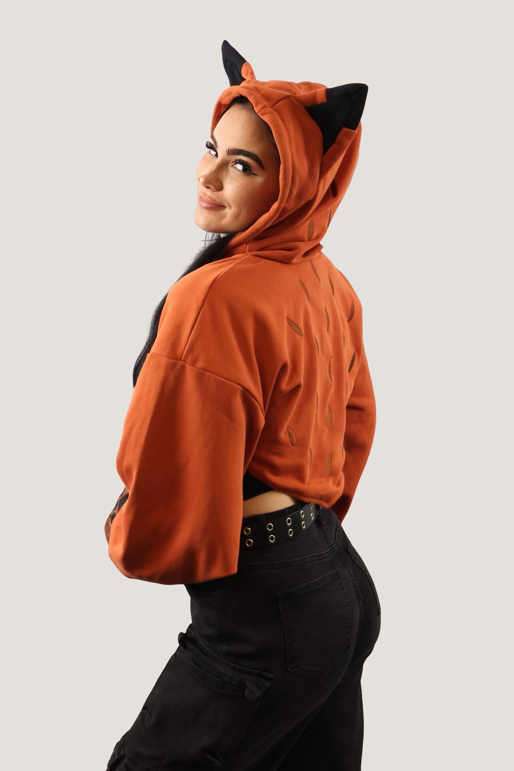 Back angled view of model wearing an orange fox-inspired hooded shrug with black ear details, showing cropped fit and hood design against a light background