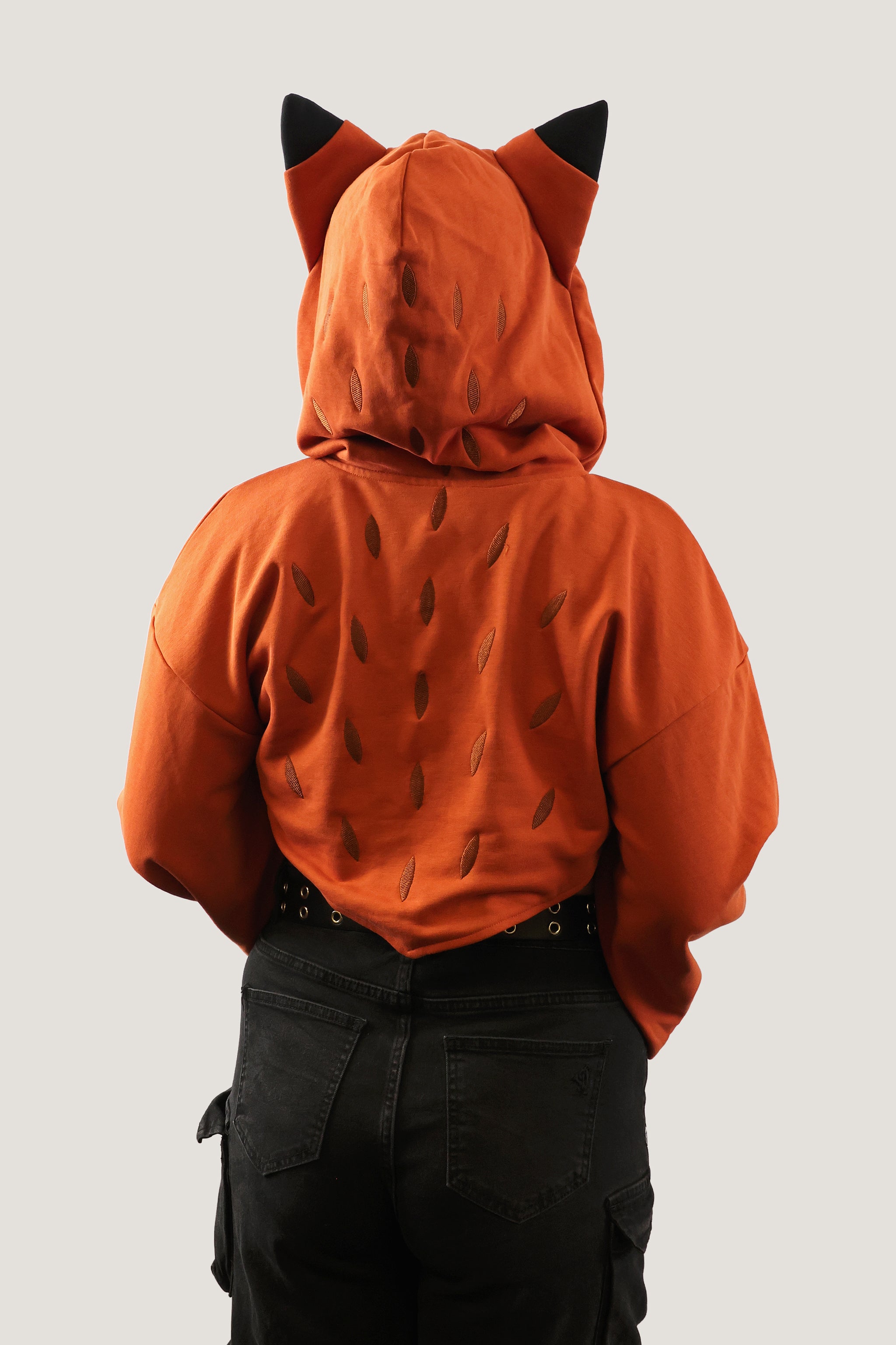 Back view of model wearing an orange fox-inspired hooded shrug with black ear details and subtle back pattern, showing cropped fit against a light background