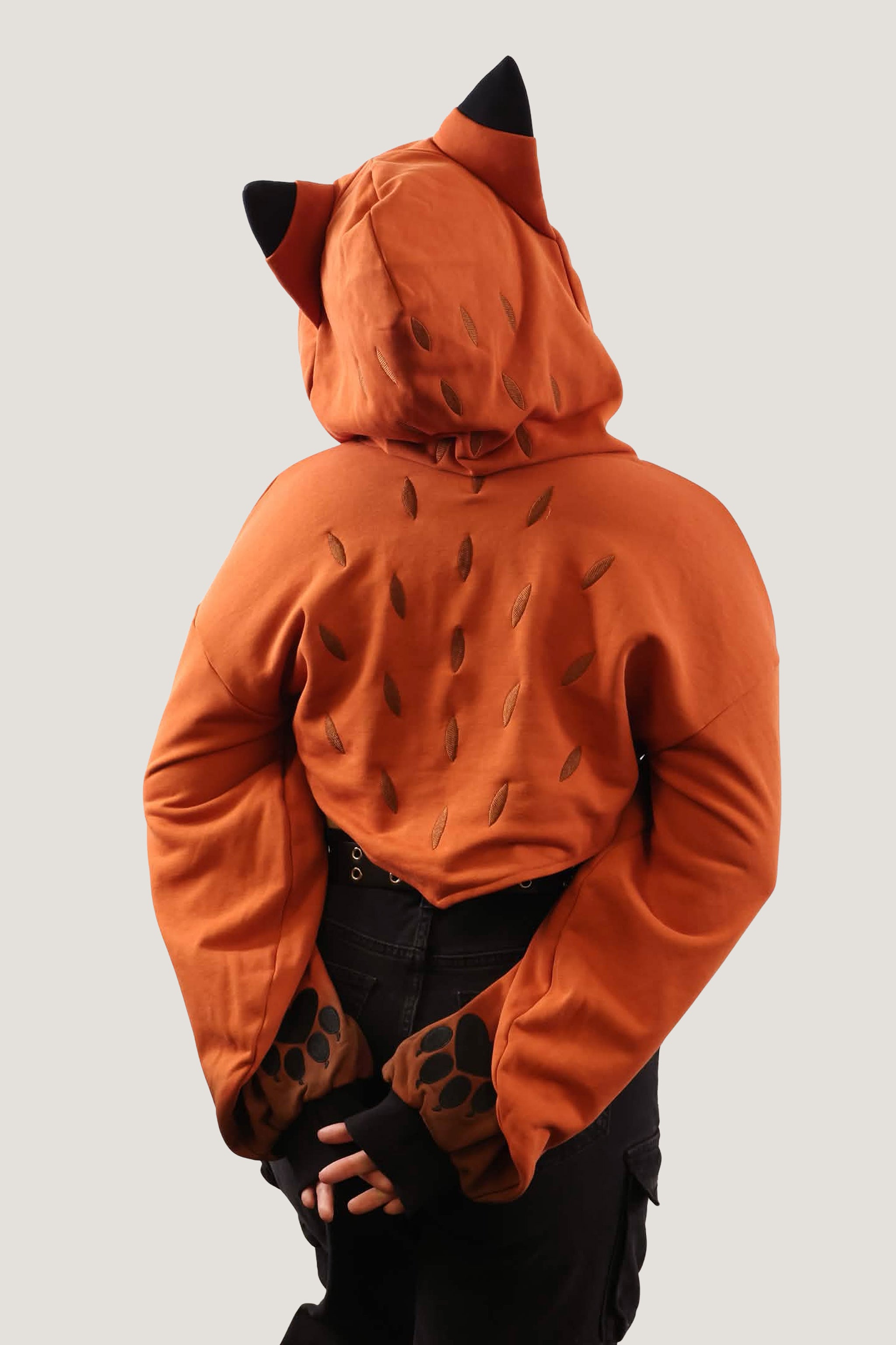 Back view of model wearing an orange fox-inspired hooded shrug with black ear details, showing back pattern and paw print sleeve cuffs against a light background