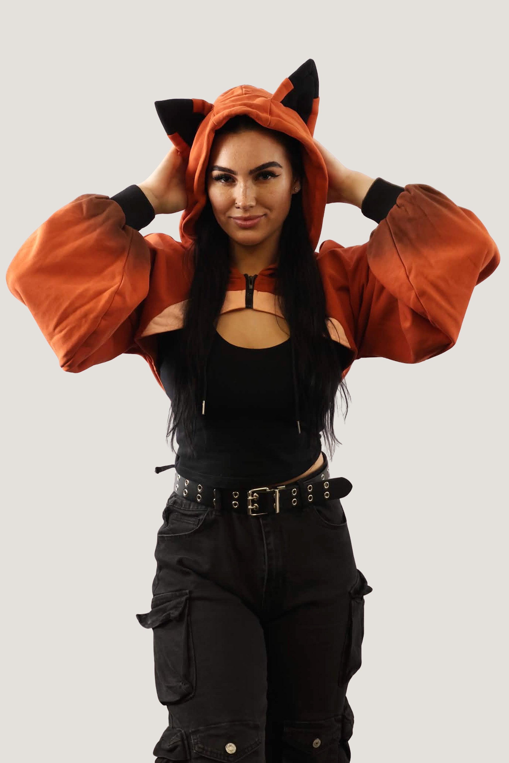 Front view of model wearing an orange fox-inspired hooded shrug with black ear details, holding the hood and showing cropped fit against a light background