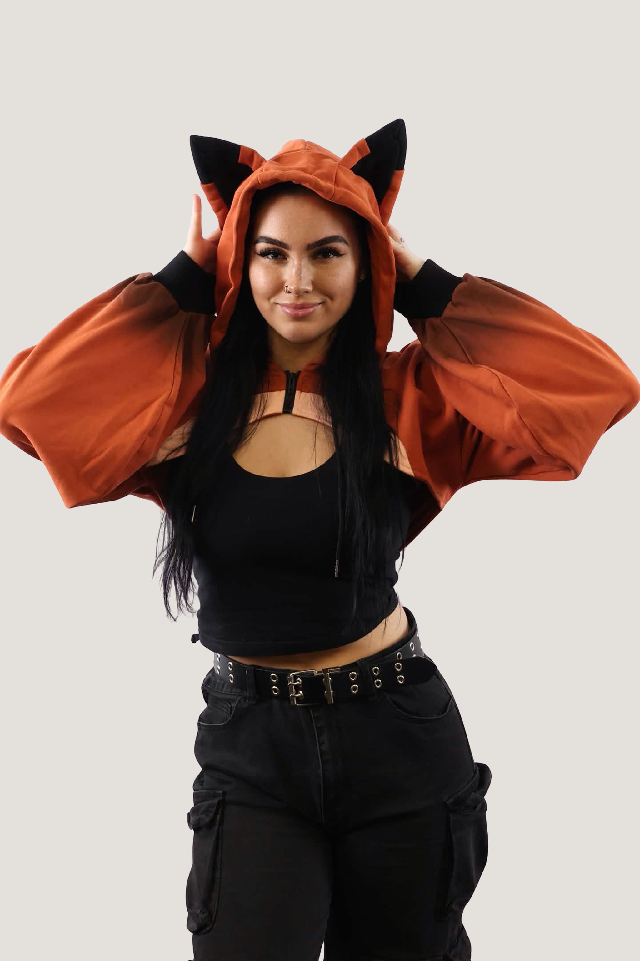 Model wearing an orange fox-inspired hooded shrug with black ear details, holding the hood and showing cropped fit against a light background