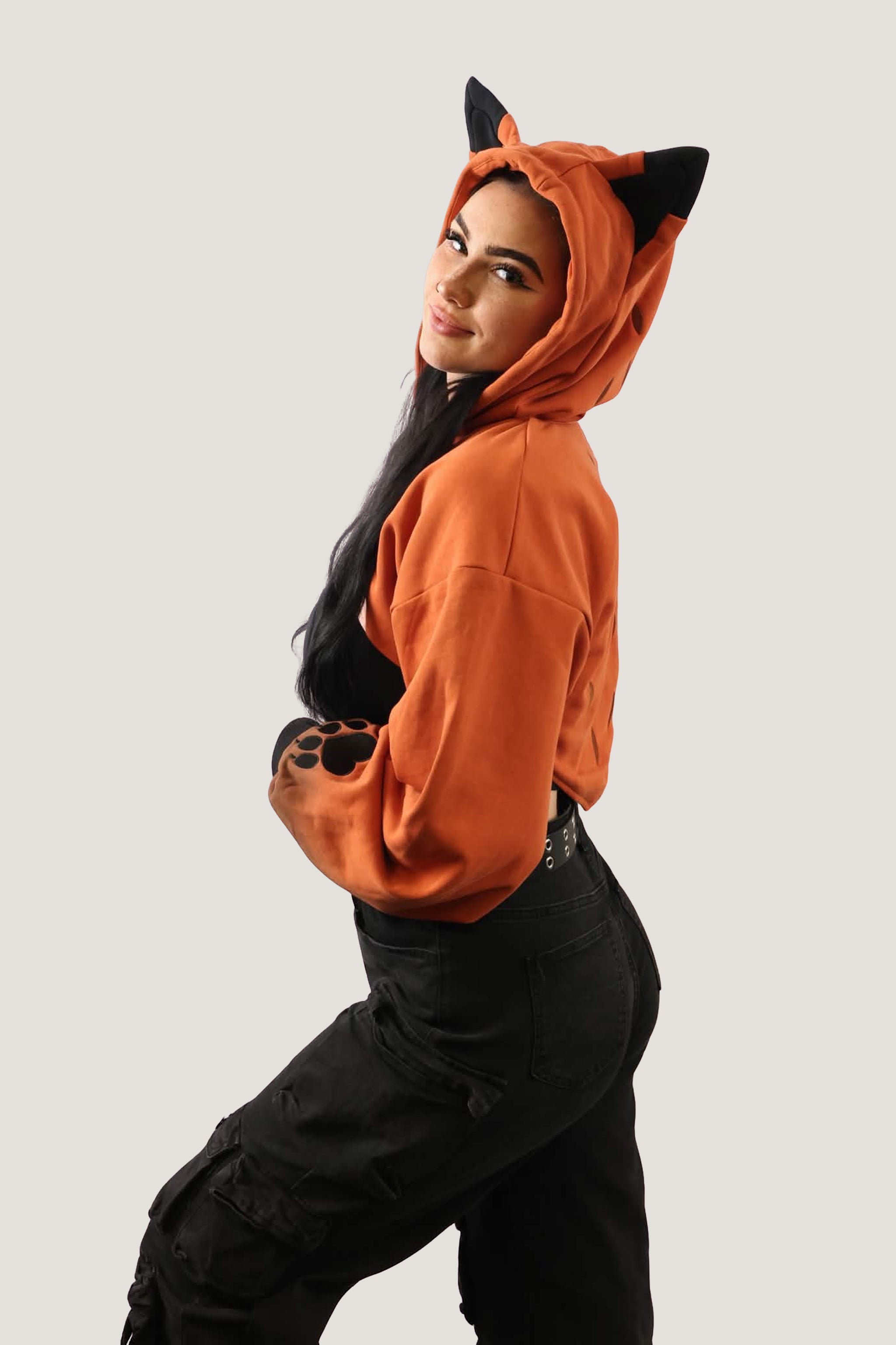 Side view of model wearing an orange fox-inspired hooded shrug with black ear details and paw print sleeve cuffs, showing cropped fit against a light background