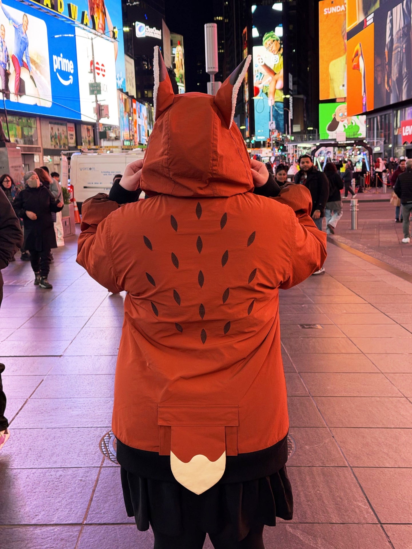 Back view of a whimsical fox-inspired jacket with ears, tail detailing, and embroidered markings photographed in Times Square at night.