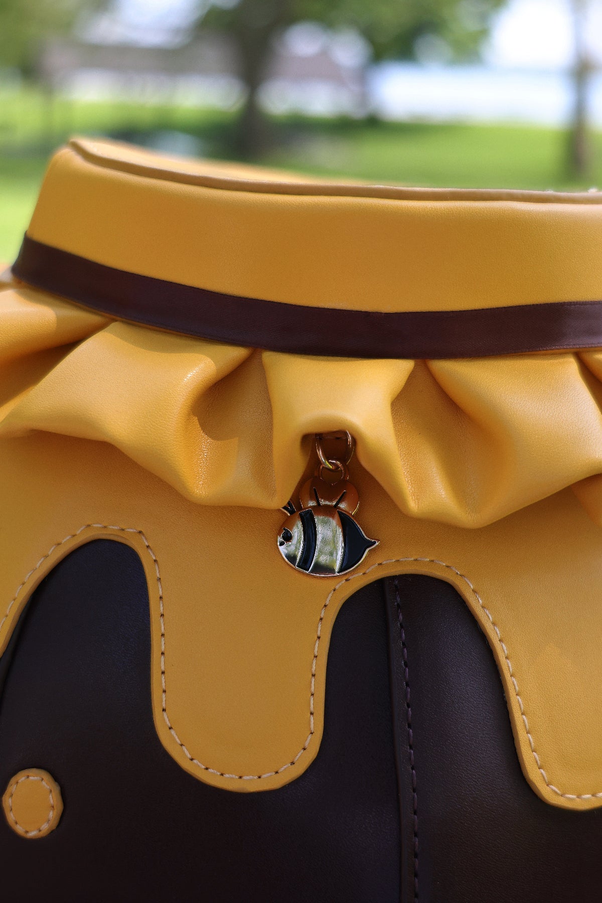Close-up detail of a honey jar shaped crossbody bag featuring honey drip accents, a brown ribbon trim, and a dangling bee zipper charm. The bag is photographed outdoors with a softly blurred green park background.