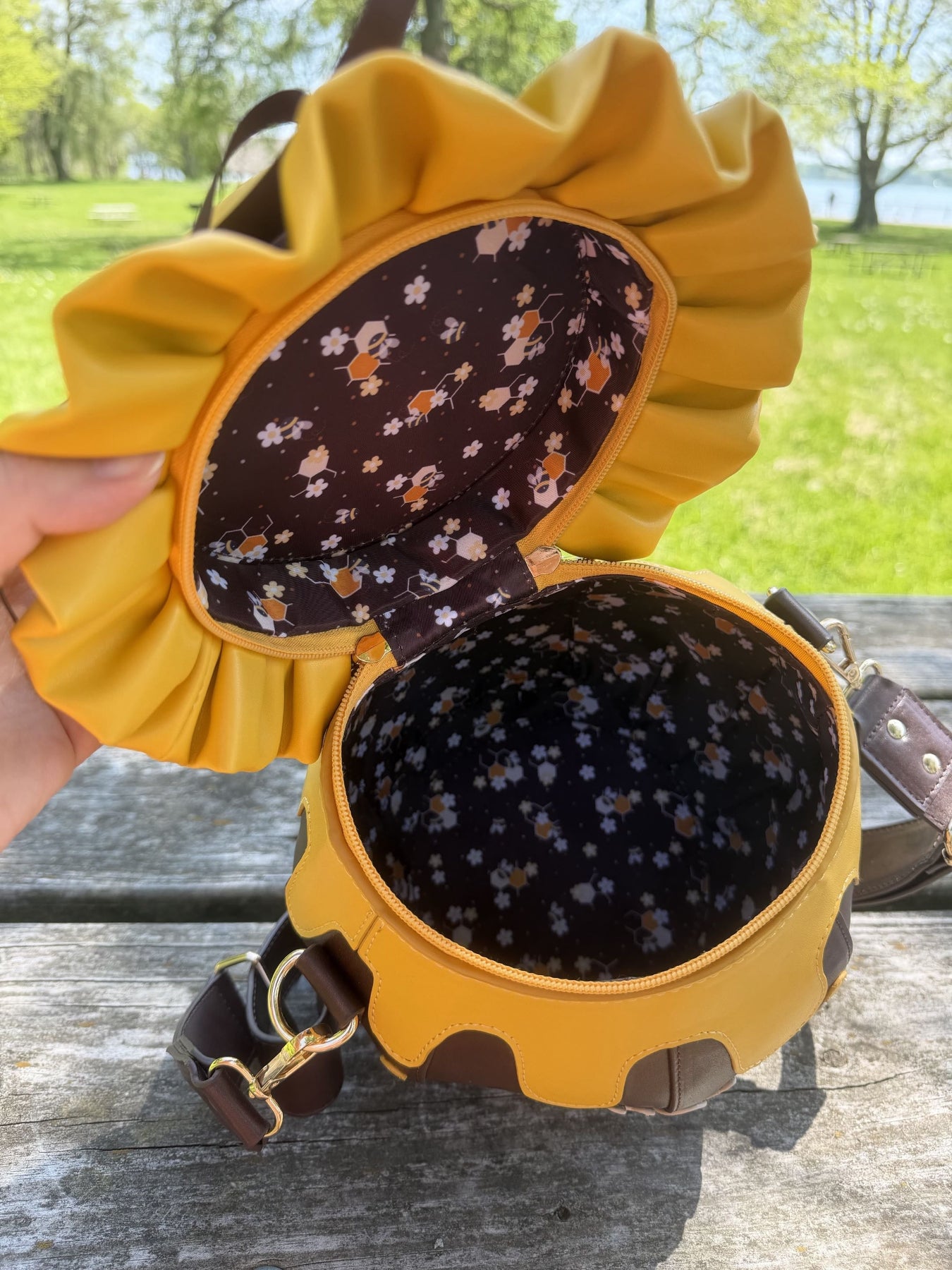Open honey jar shaped crossbody bag showing its spacious interior and custom bee floral print lining. The bag features honey drip details, a ruffled lid, and a brown shoulder strap, photographed outdoors on a wooden picnic table.