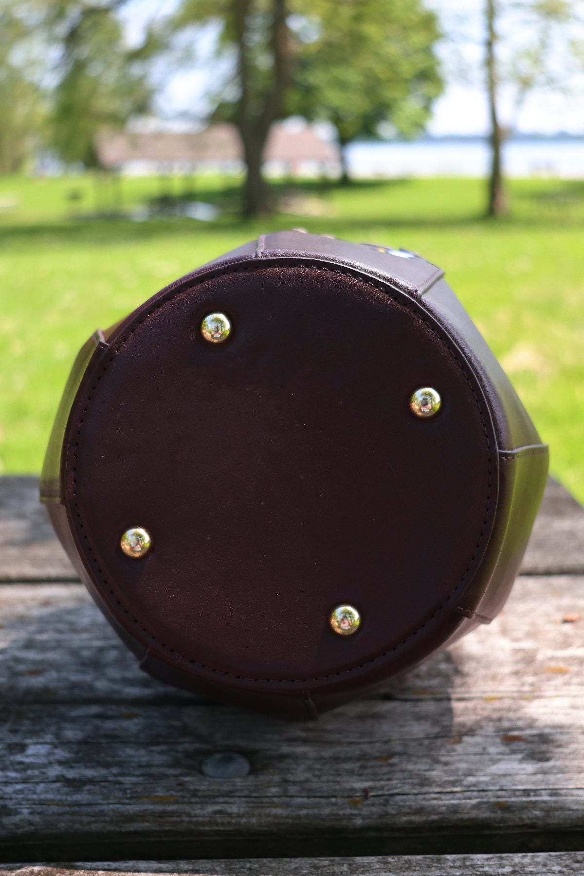 Bottom view of a honey jar shaped crossbody bag showing the round brown base, protective metal feet, and structured construction. The bag is photographed outdoors on a wooden picnic table with a grassy park background.