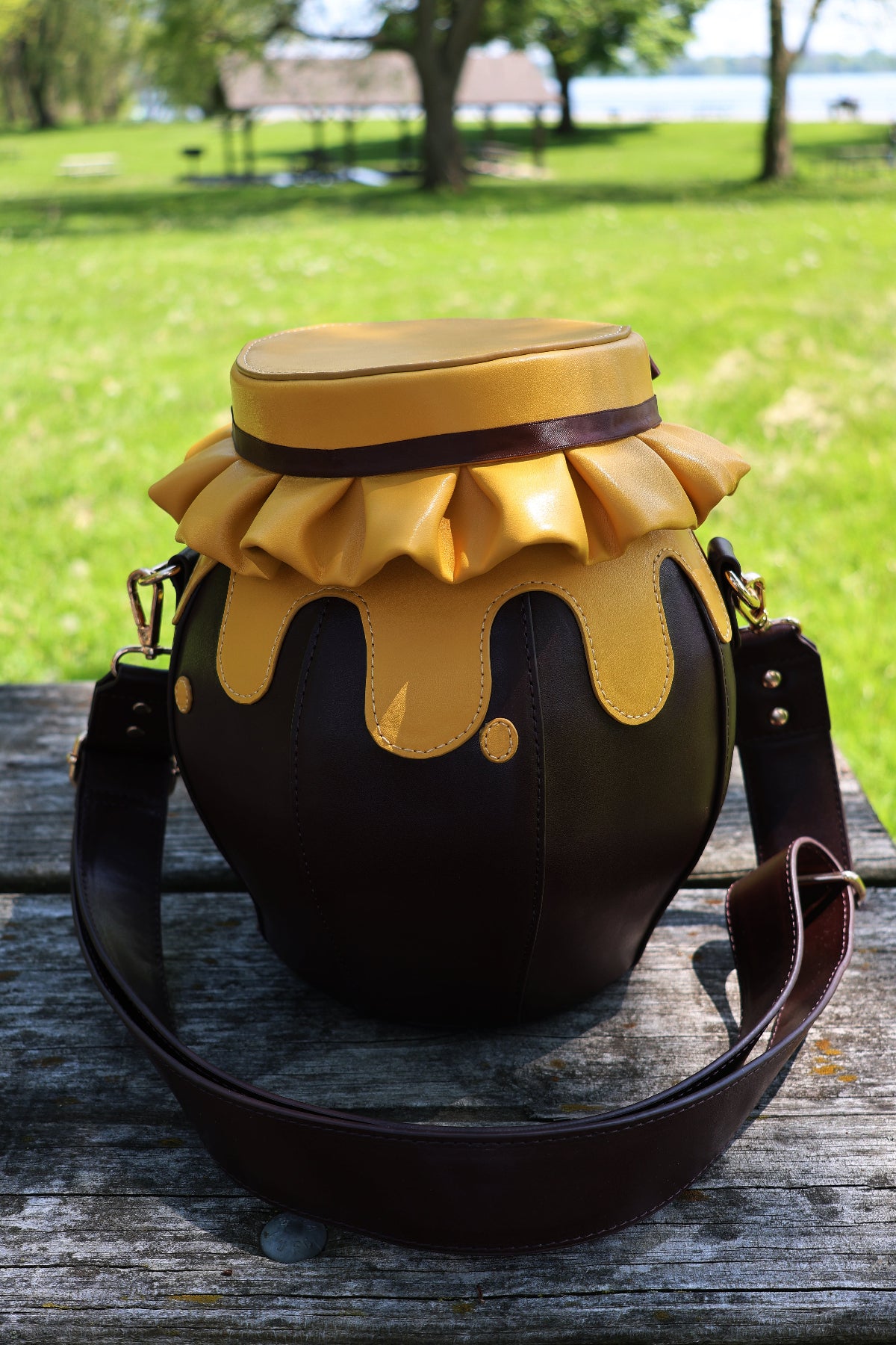 Back view of the Honeypot Crossbody Bag featuring a honey jar silhouette, dripping honey details, and an adjustable brown crossbody strap, photographed outdoors on a wooden picnic table.