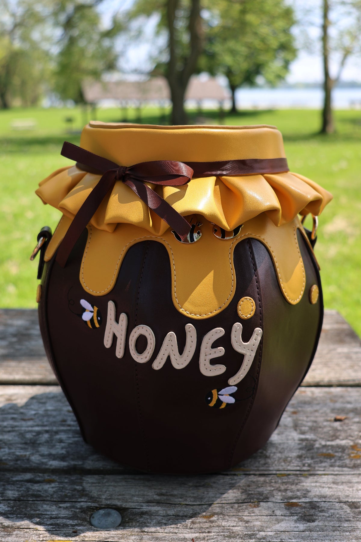 Front view of the Honeypot Crossbody Bag shaped like a honey jar, featuring a dripping honey lid, brown ribbon bow, embroidered bees, and “HONEY” lettering, displayed outdoors on a wooden picnic table.