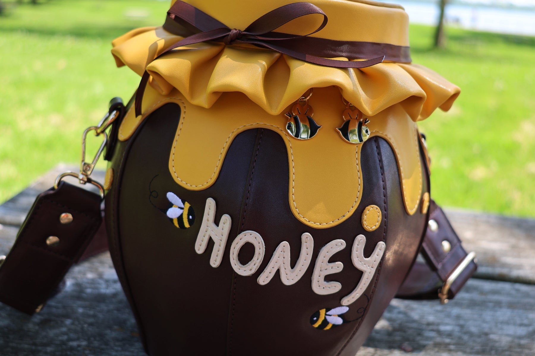 Close-up of the Honeypot Crossbody Bag featuring a honey jar-inspired design with dripping honey details, embroidered bees, bee-shaped zipper charms, and "Honey" lettering on the front.
