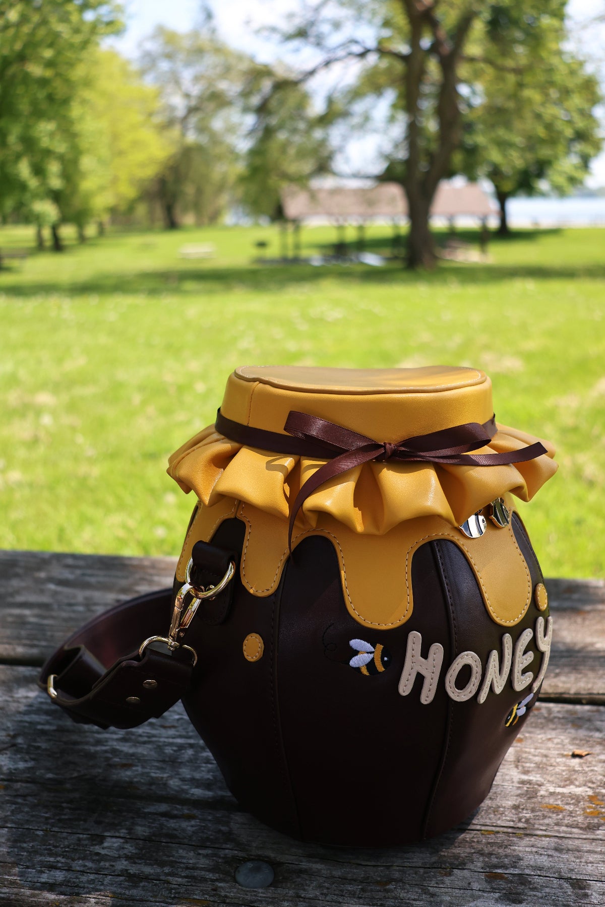 Honeypot Crossbody Bag shaped like a honey jar with dripping honey details, bee accents, and a ribbon-tied lid, displayed outdoors on a wooden picnic table in a sunny park setting.