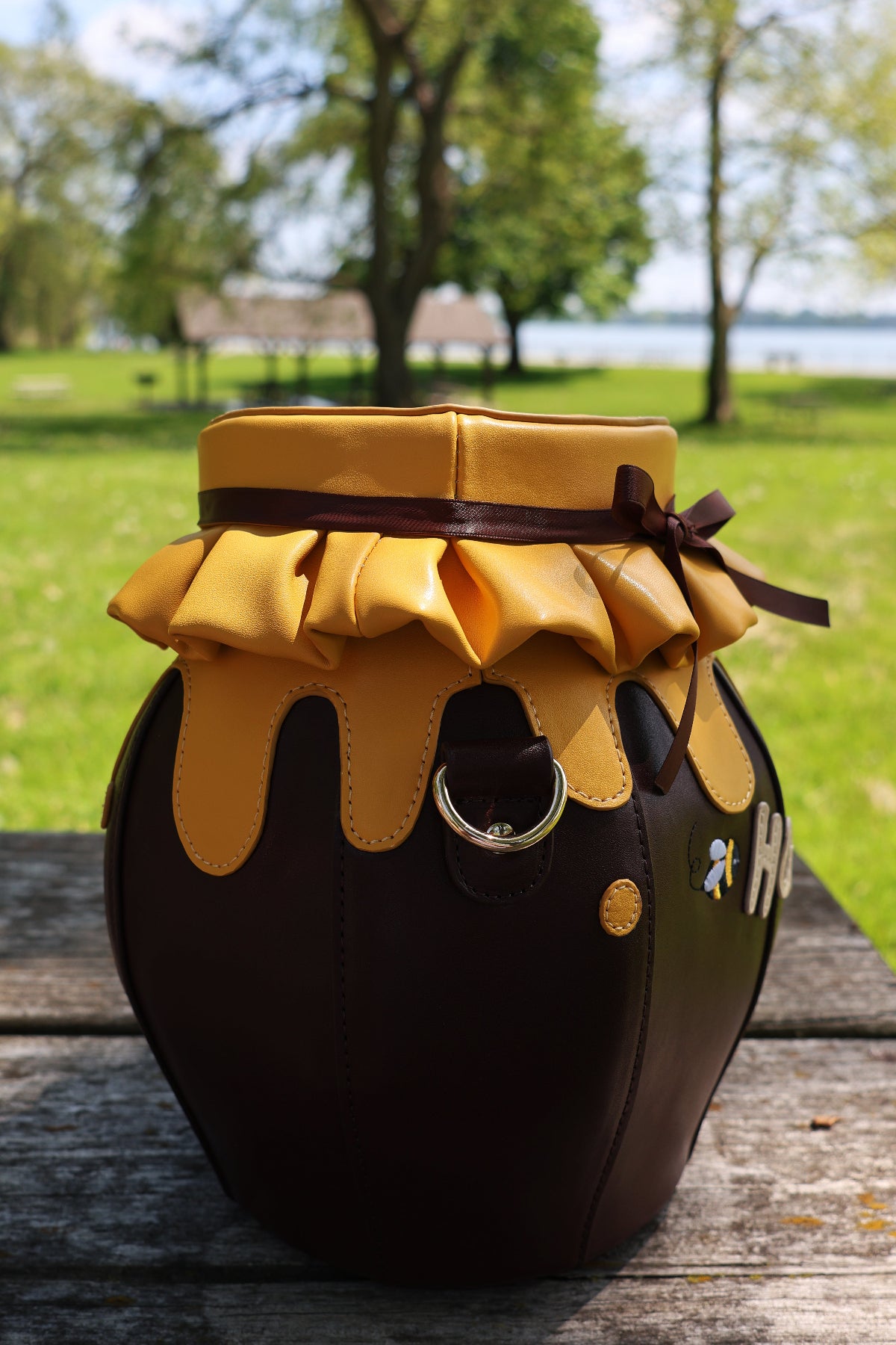 Side view of the Honeypot Crossbody Bag featuring a honey jar shape, dripping honey lid detail, brown ribbon accent, and bee embellishments, photographed outdoors on a wooden picnic table.