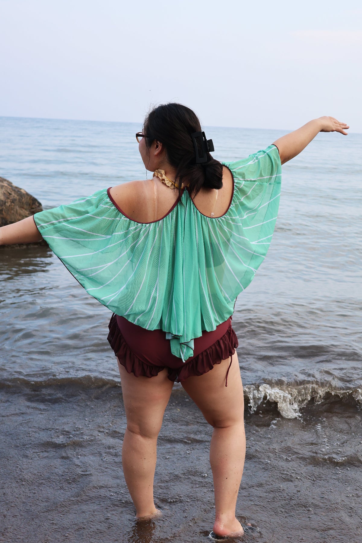 Back view of the Nyahallo Luna Moth Swimsuit at the beach, featuring a flowing mint green moth wing-inspired cape and burgundy ruffle bottoms.