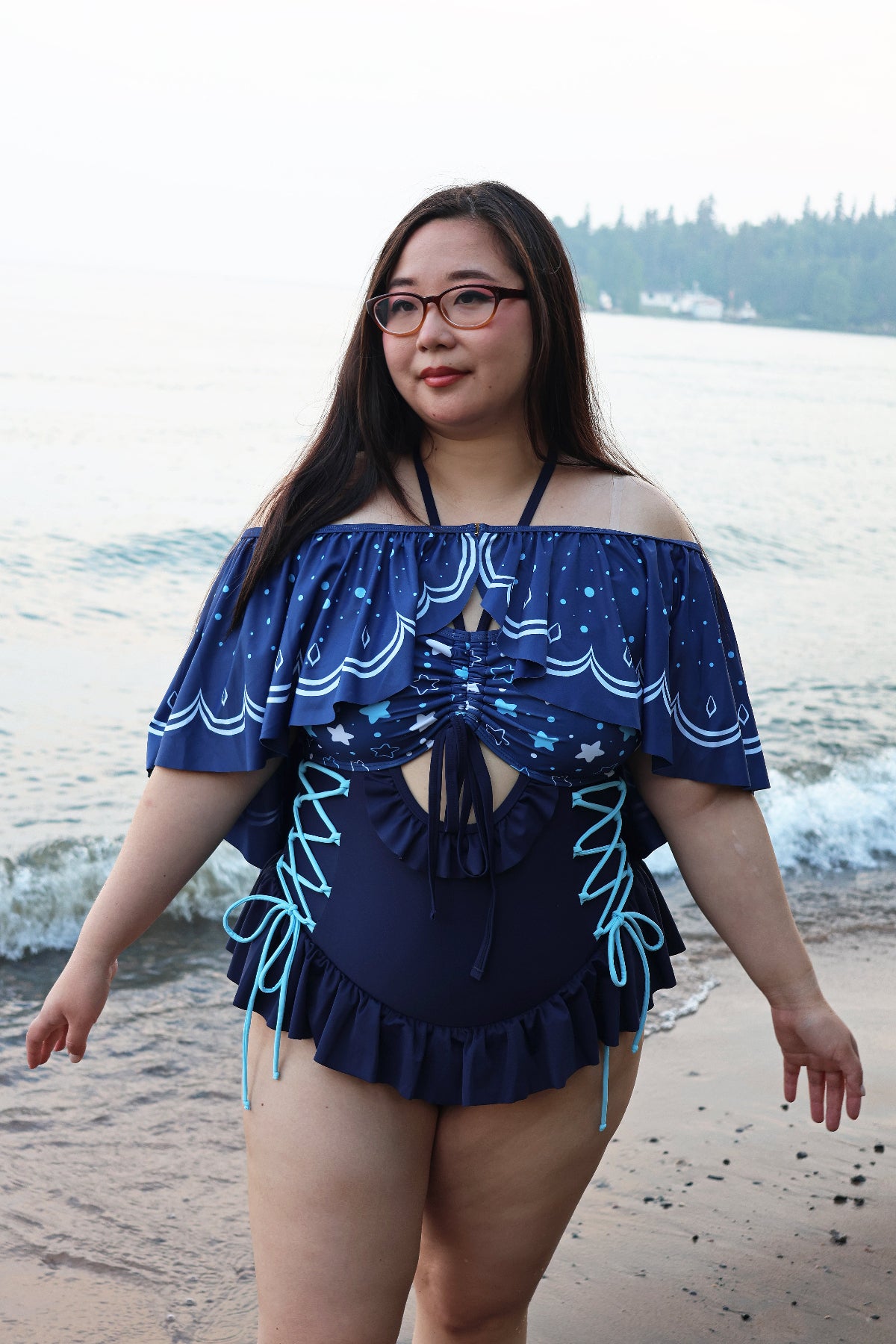 Model wearing the Nyahallo Moon Jellyfish Swimsuit with the detachable moon jellyfish-inspired cape, featuring a star-patterned bust, lace-up sides, and flowing cape while walking along the beach.