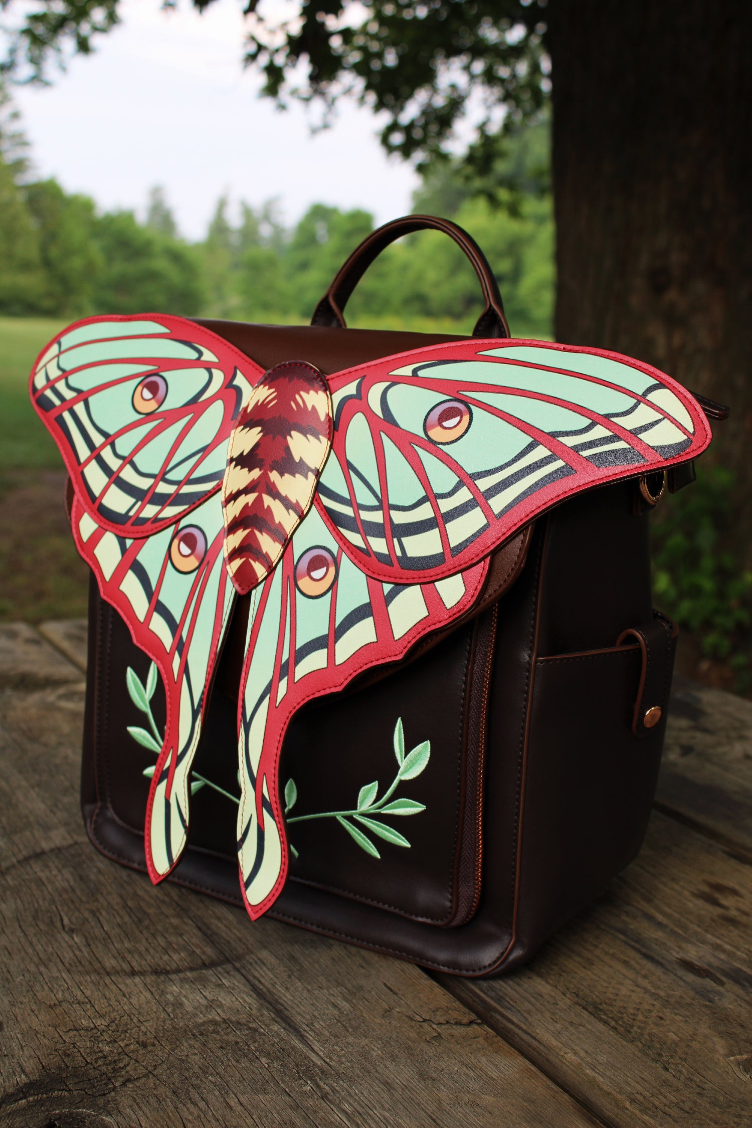 Front view of a brown backpack with large moth wing flap, photographed on a wooden table outdoors with greenery in the background.