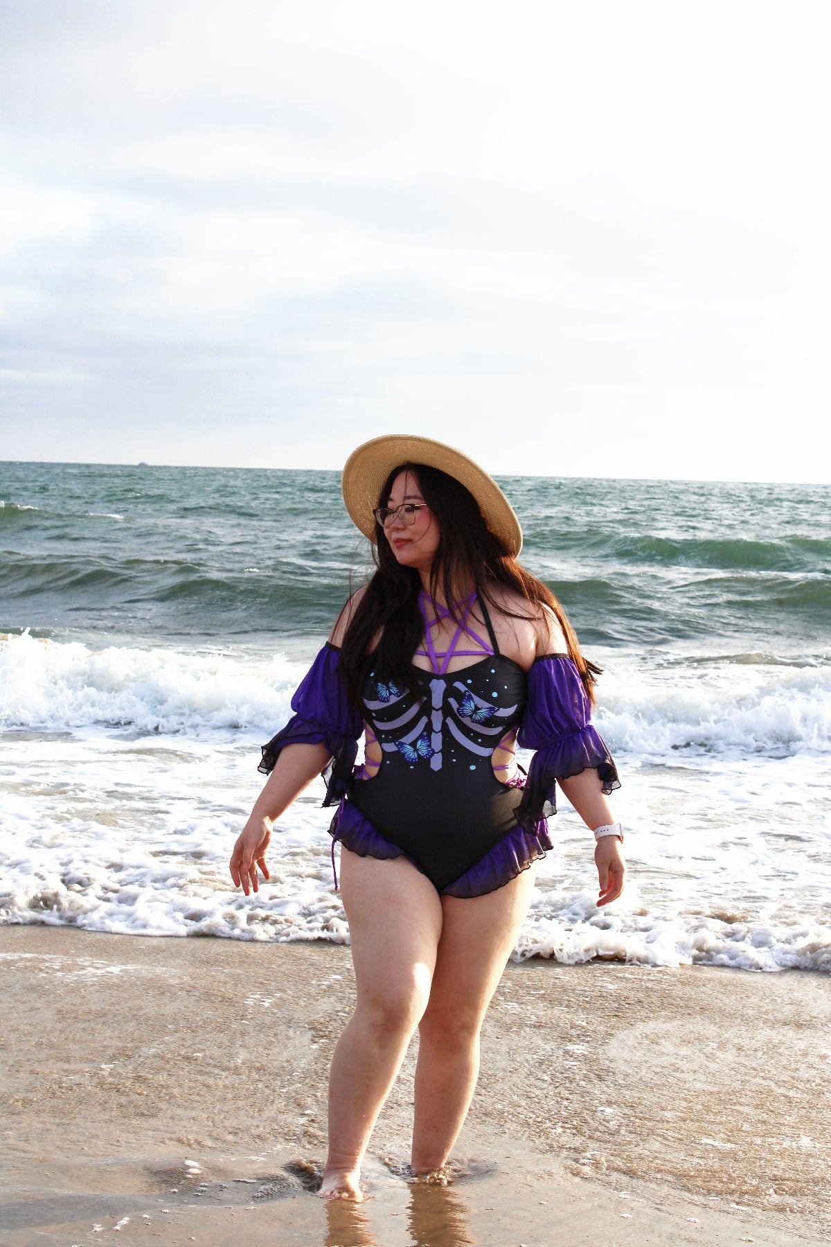 Model walking along the beach wearing the Nyahallo Necromancer One-Piece Swimsuit with sheer purple sleeves, a skeletal butterfly-inspired print, and lace-up neckline.