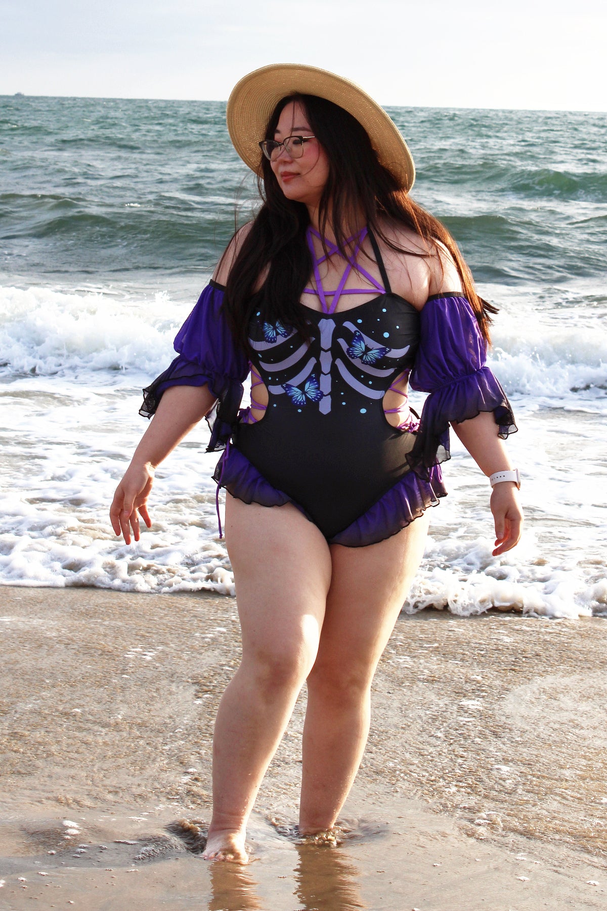 Model walking along the shoreline wearing the Nyahallo Necromancer One-Piece Swimsuit with a skeletal butterfly print, sheer purple sleeves, and lace-up neckline.