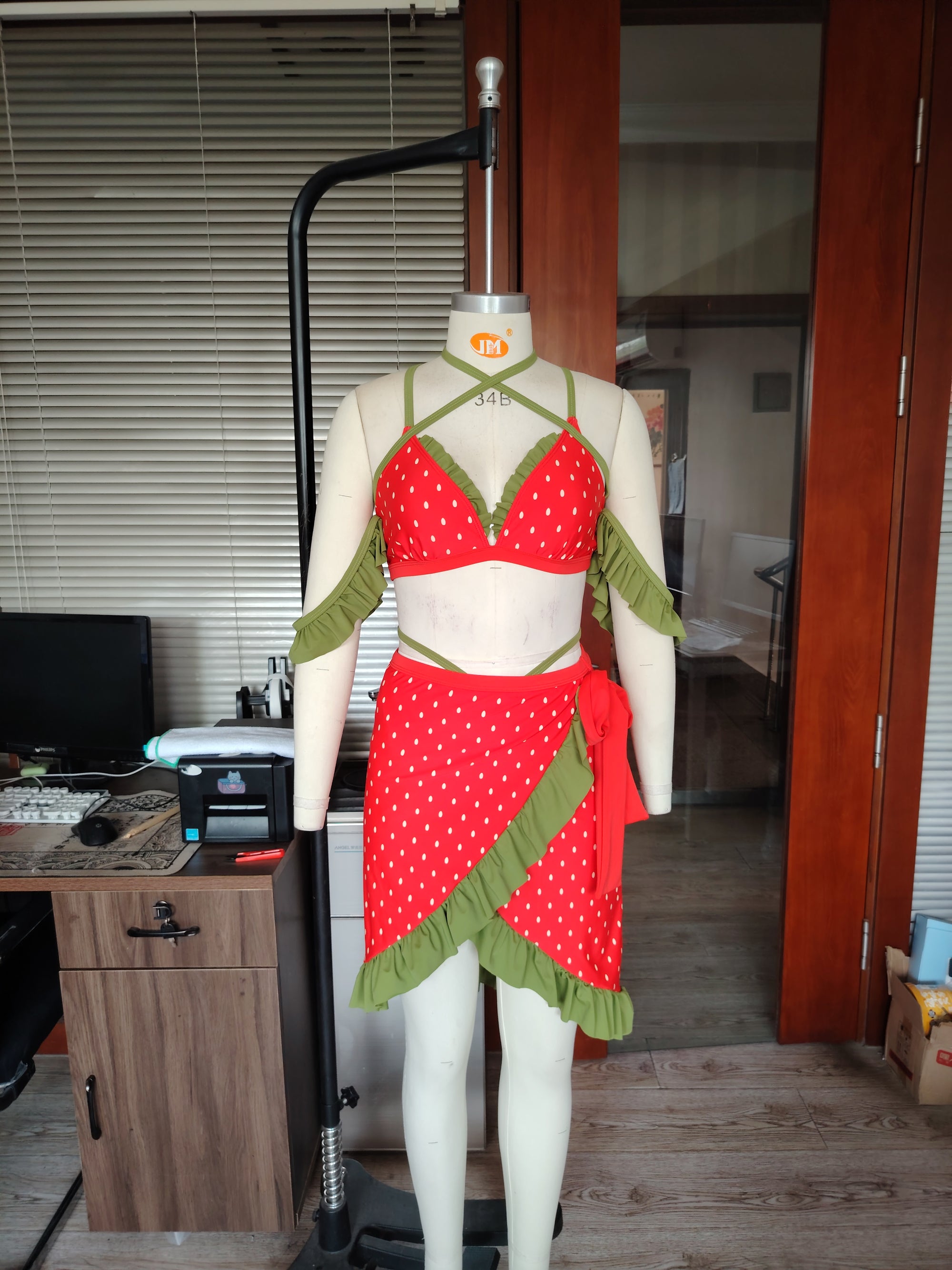 Mannequin wearing a red and green strawberry themed bikini with wrap skirt in an indoor setting.