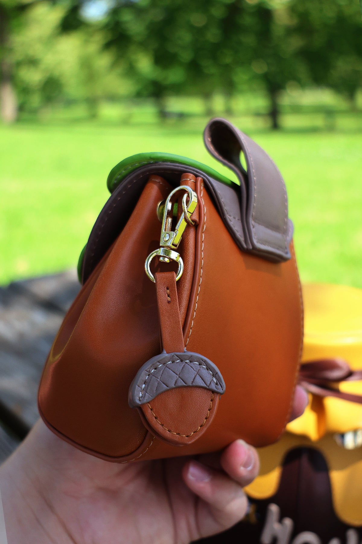 Side view of a small brown oak leaf pochette featuring a detachable acorn charm and gold-tone hardware, photographed outdoors in a park.