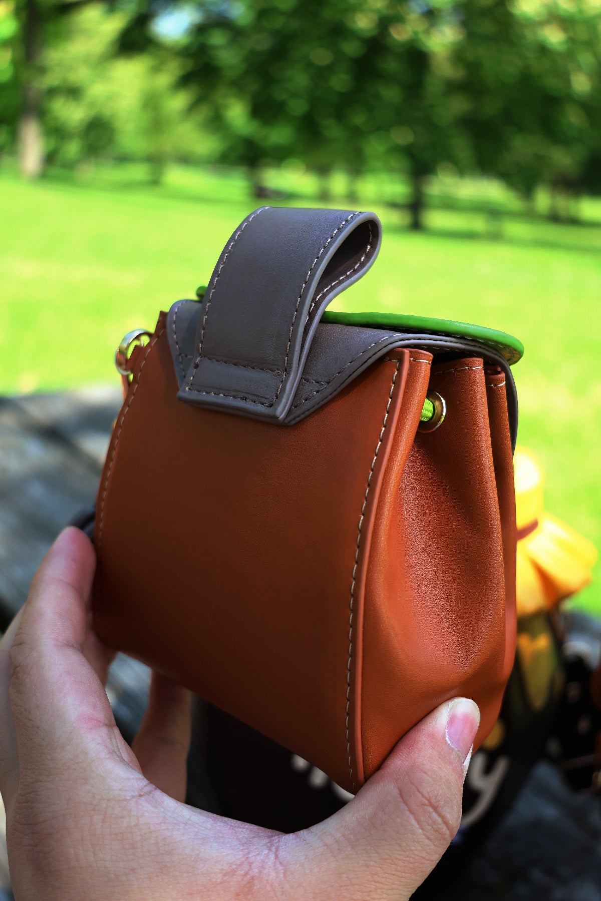 Back view of a small brown oak leaf pochette showing the smooth faux leather exterior and structured silhouette, photographed outdoors in a park.