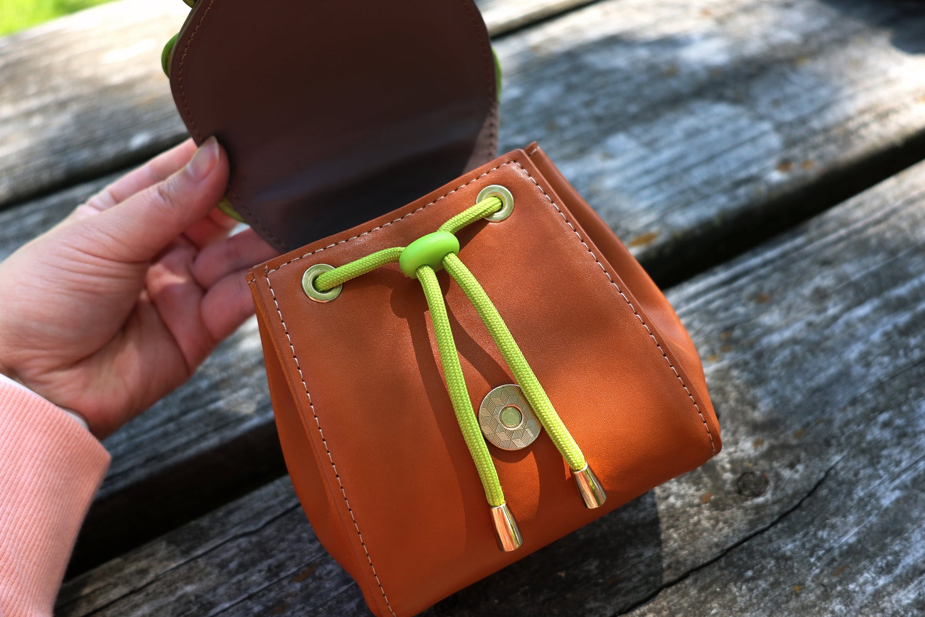 Open brown faux leather pochette featuring a green drawstring closure and roomy pouch interior, shown in natural outdoor lighting.