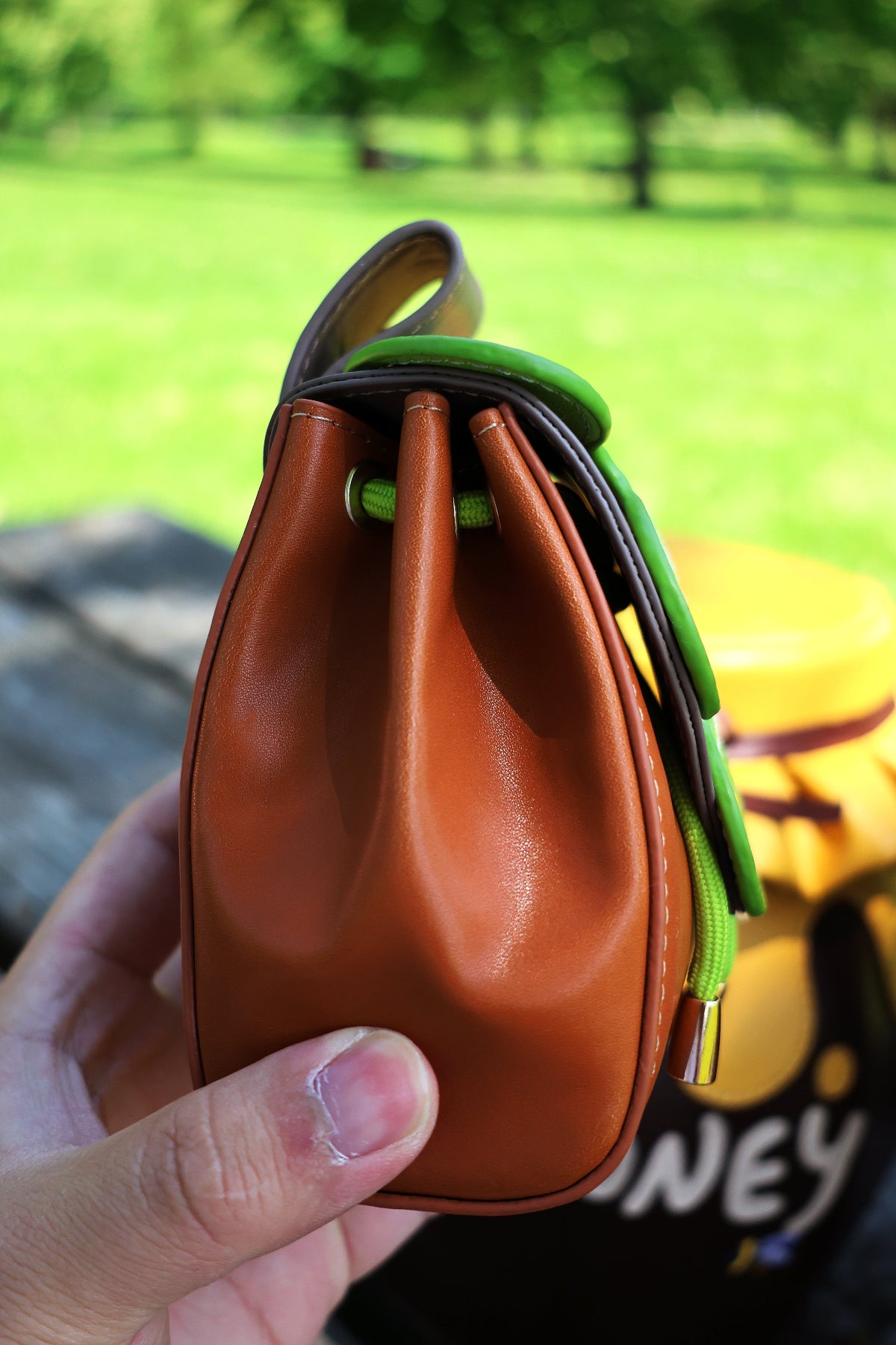 Side view of a small brown oak leaf pochette showing the gathered silhouette and drawstring closure, photographed outdoors in a grassy park.