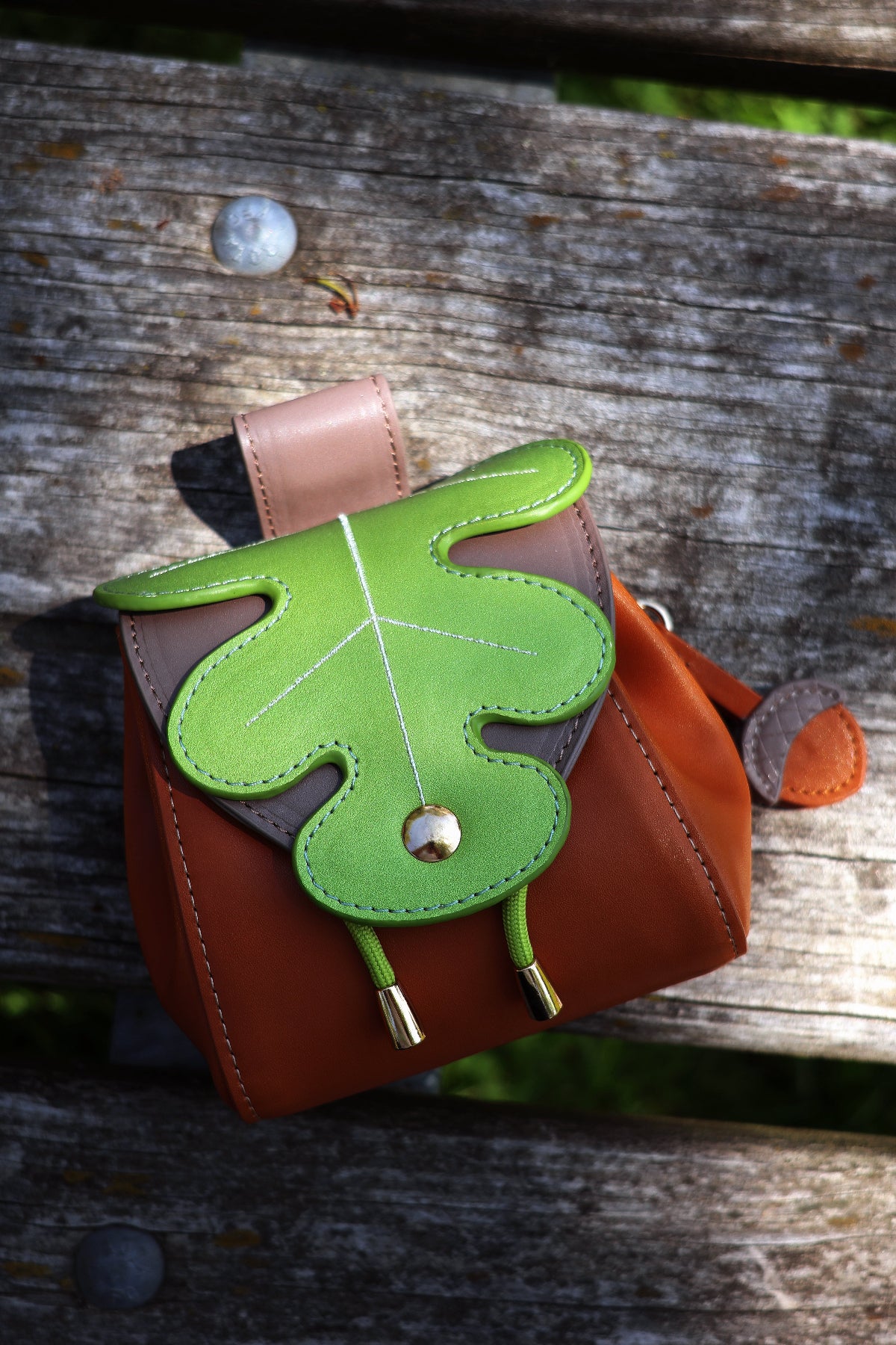 Top-down view of a small brown oak leaf pochette with a green leaf-shaped flap and acorn charm, displayed on a weathered wooden bench outdoors.